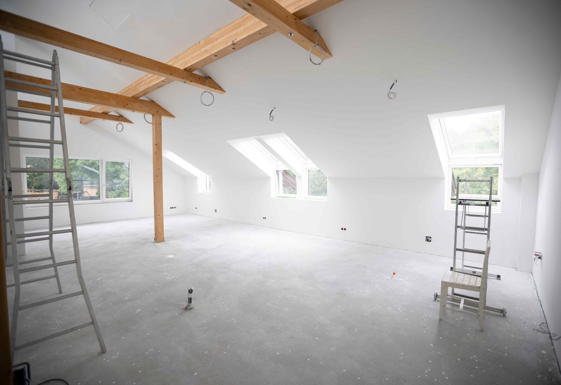 Interior of a white, unfinished attic space with wooden beams and skylight. Two ladders are in the room.