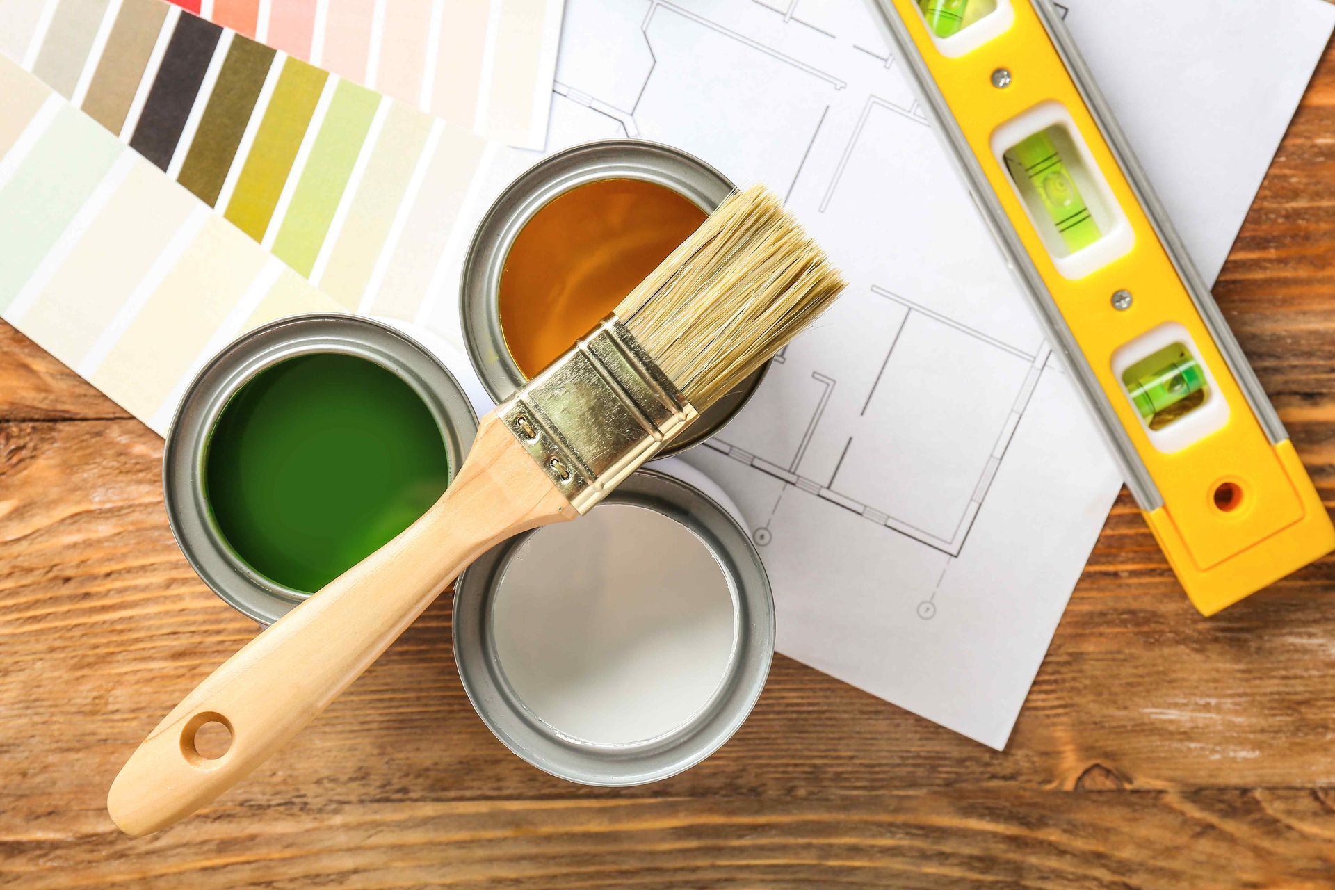 Paint cans, paintbrush, color swatches, level, and blueprints on a wooden surface.