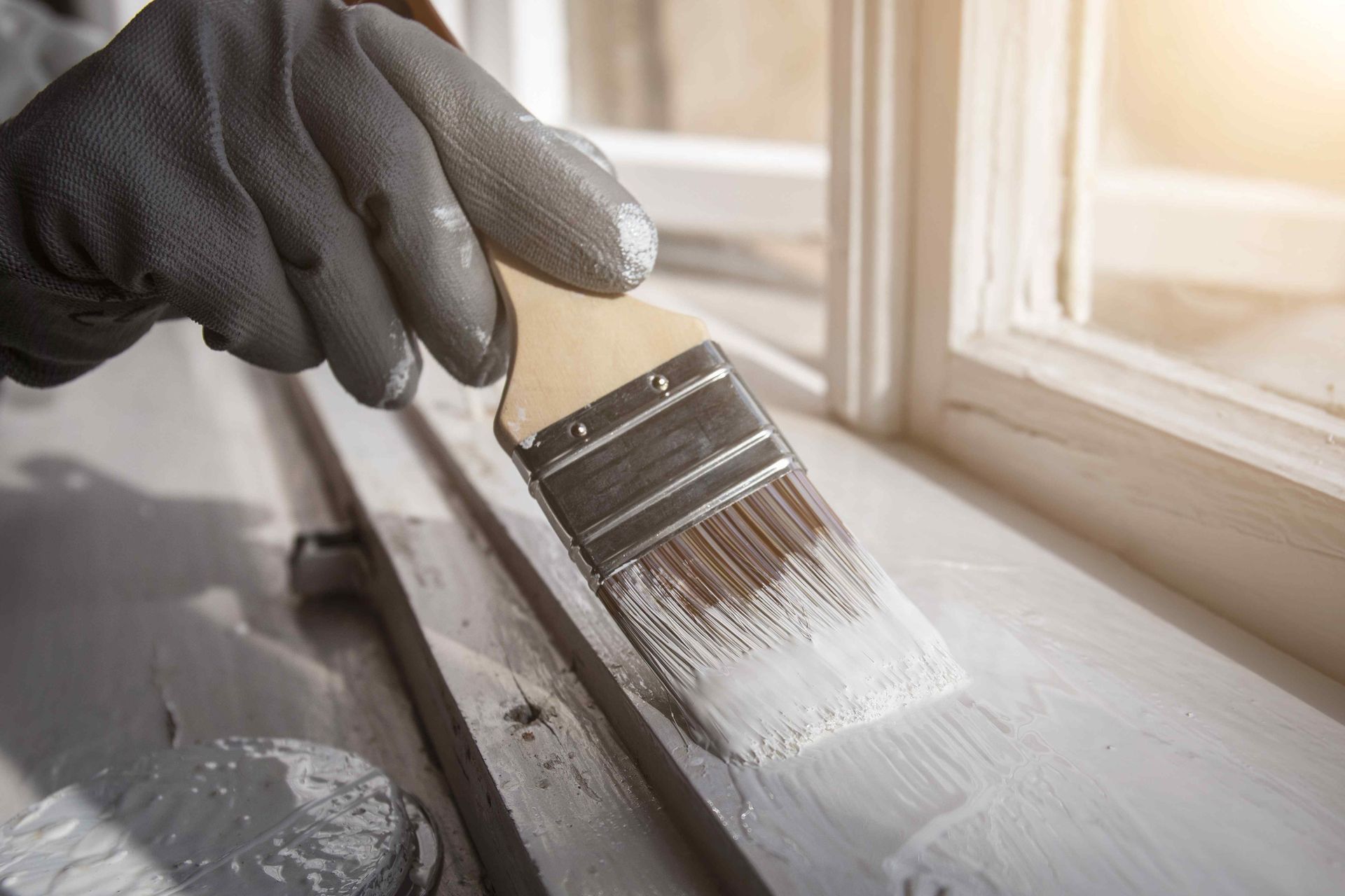 Gloved hand paints white paint onto a wooden window frame with a paintbrush.
