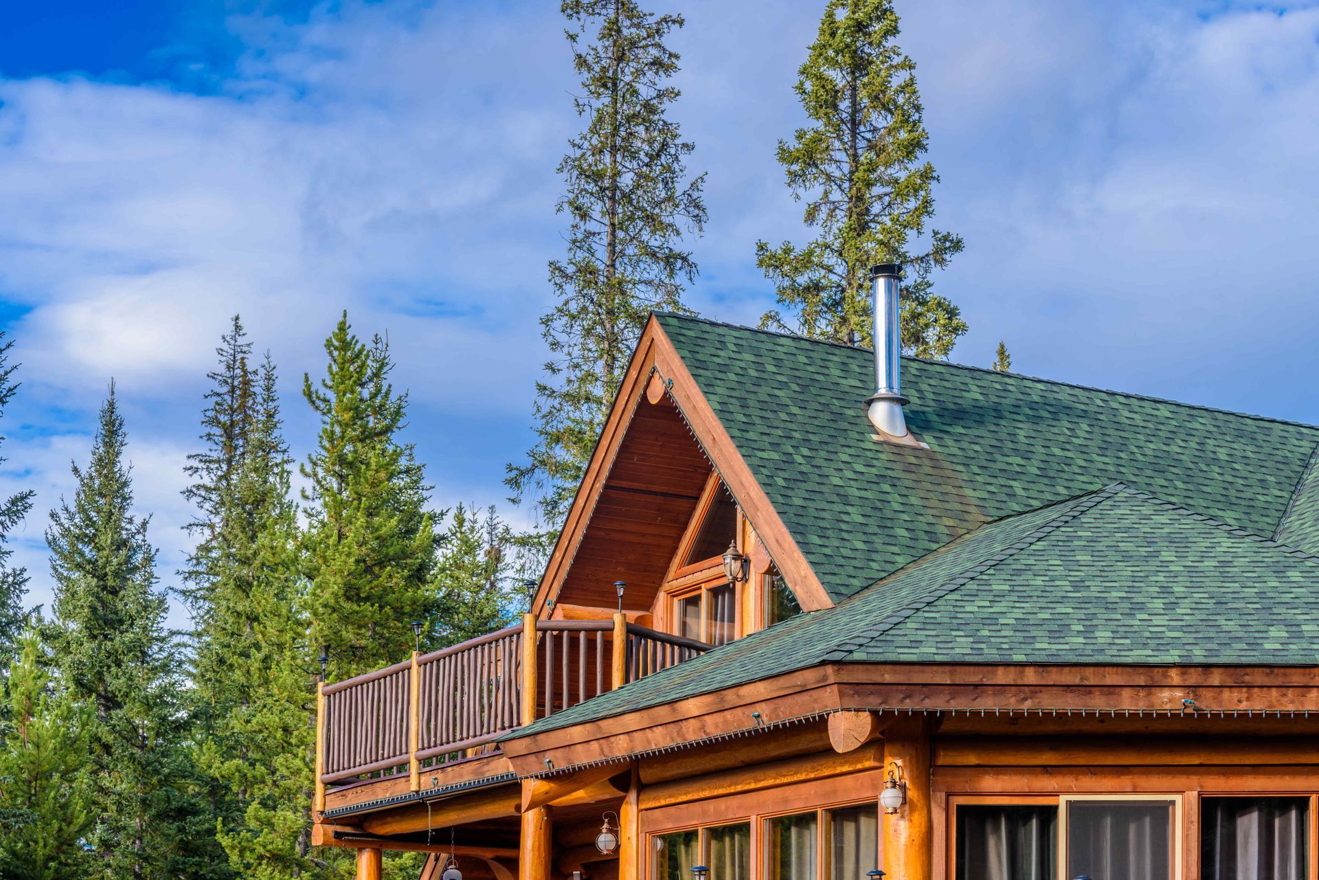 Wooden cabin with green roof, balcony, and chimney, set against a backdrop of trees and blue sky.