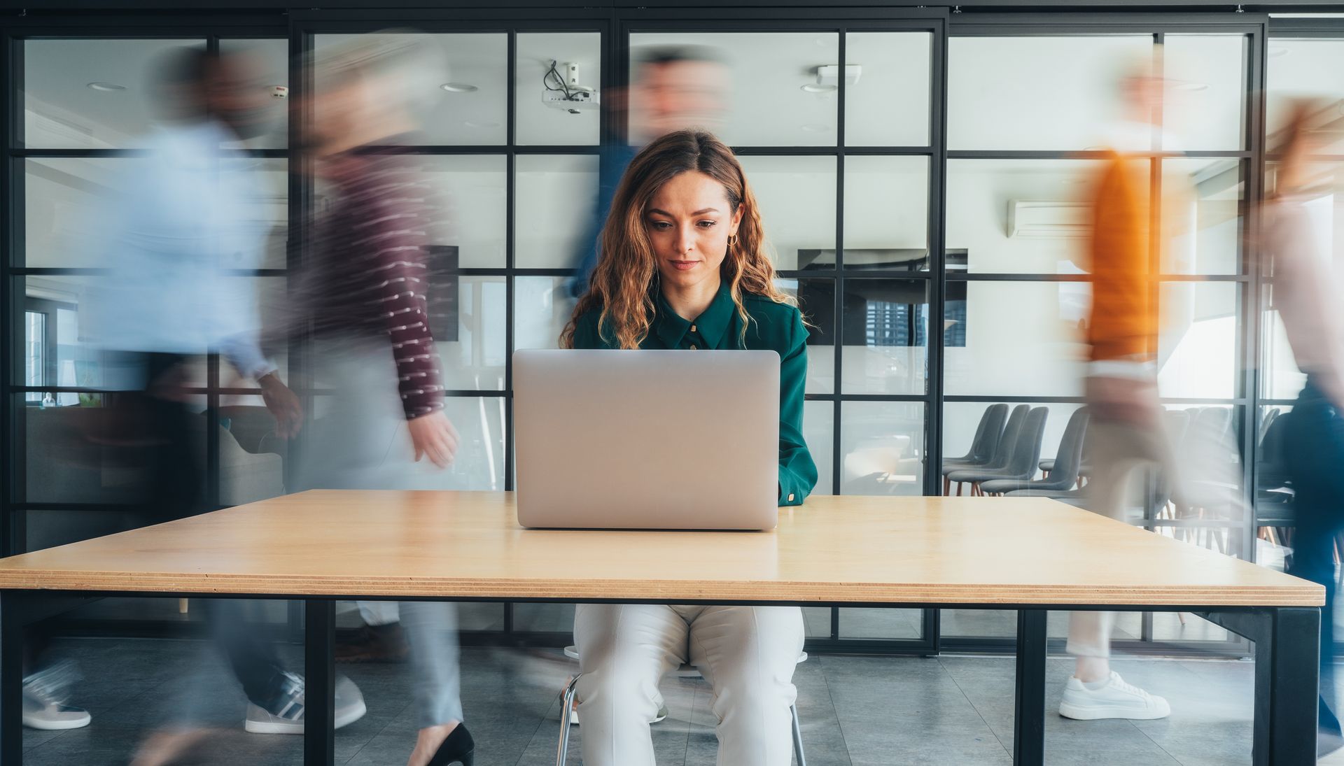 Woman working on laptop at desk, blurred people walking past in modern office setting.