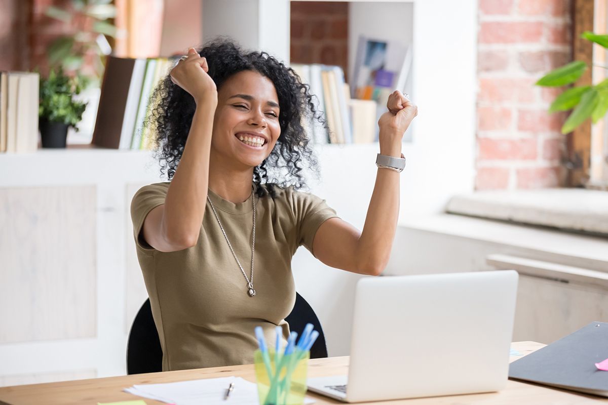 Woman at desk, celebrating, arms raised. Laptop and office supplies.