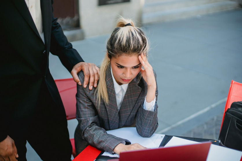Tired medical worker in scrubs sits head in hands; others rush past. Light-filled room with chairs.