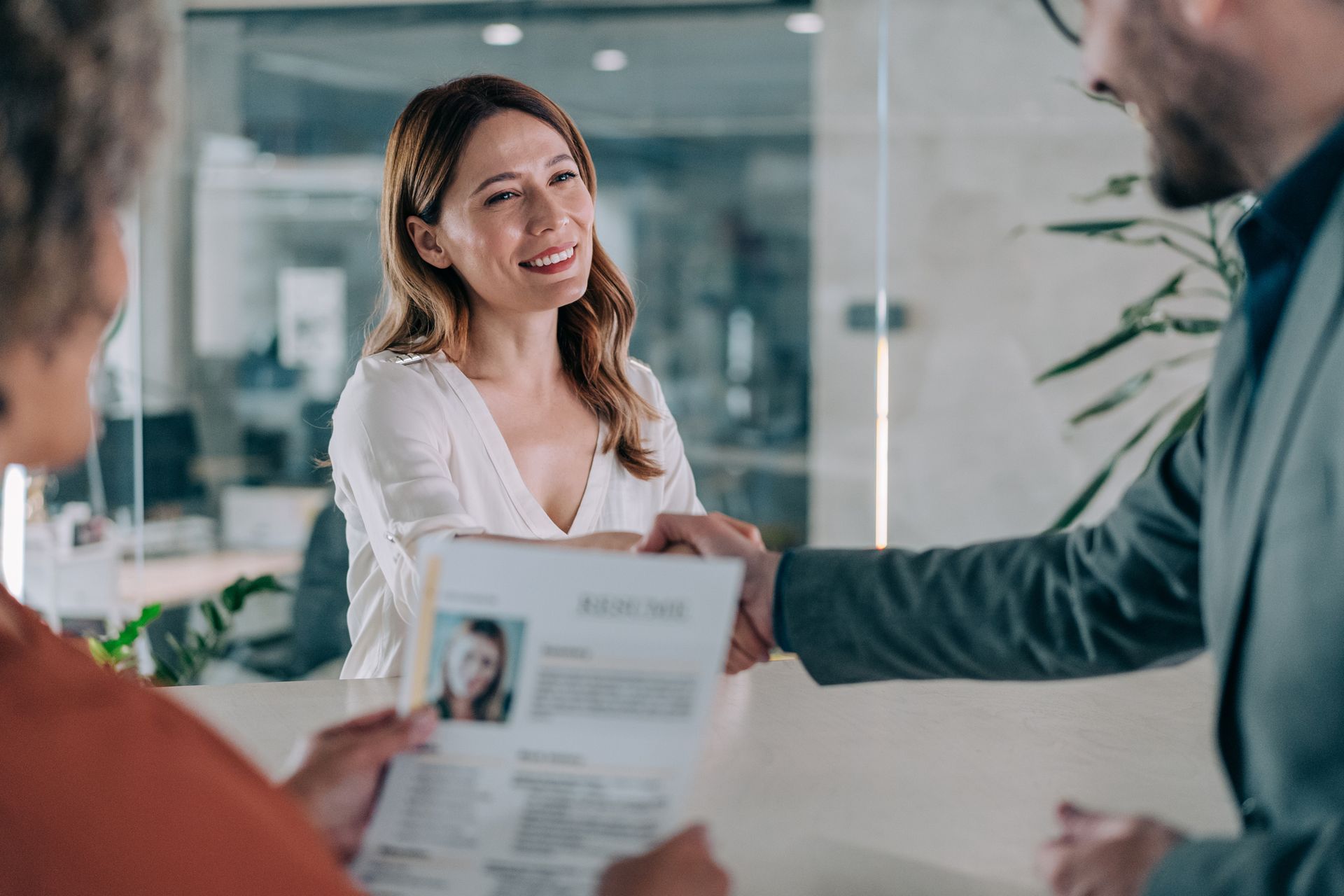 Woman smiling, handing resume to person behind desk; office setting.