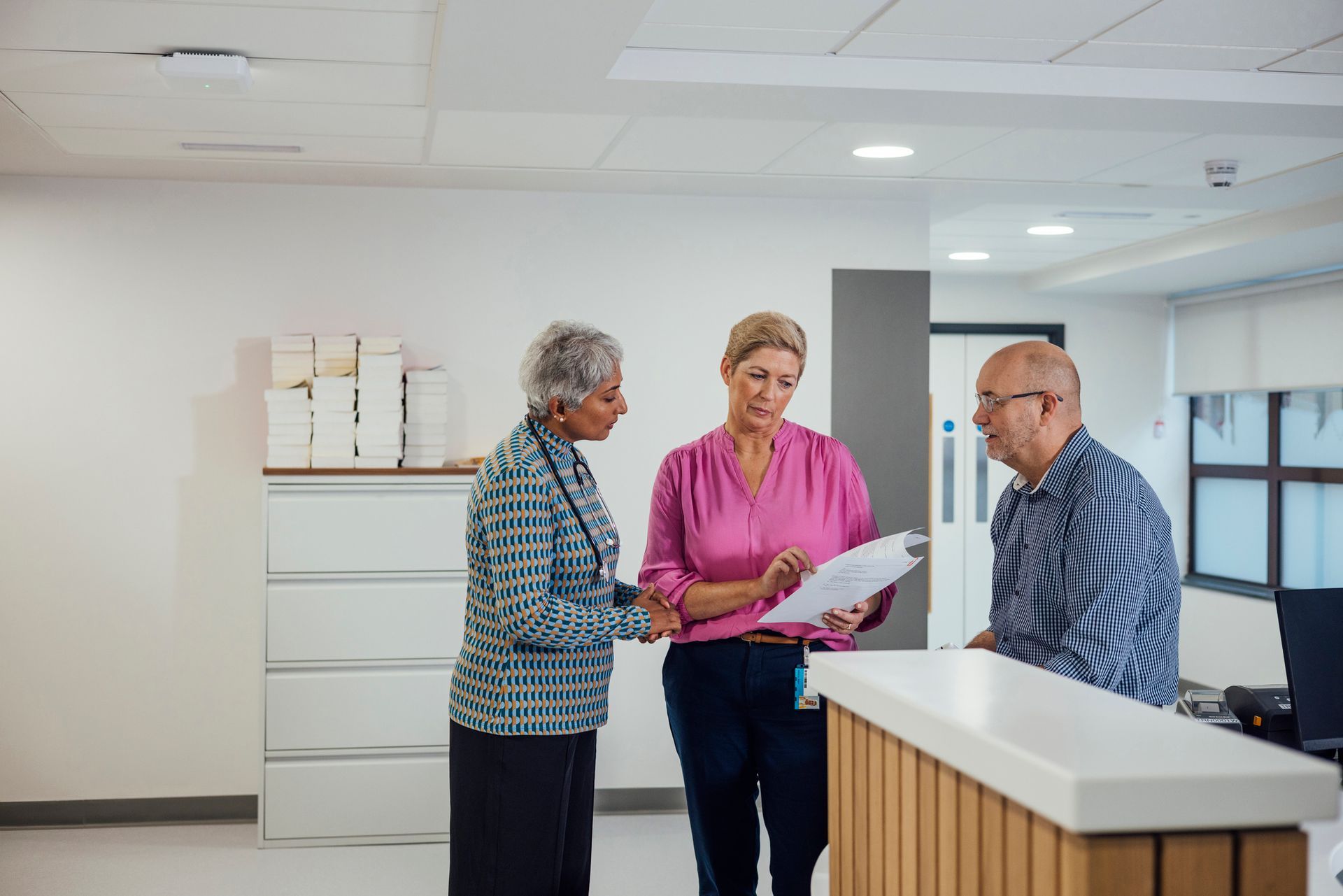 Three people at a reception desk; one person examines paperwork, another looks on, and the third stands by.