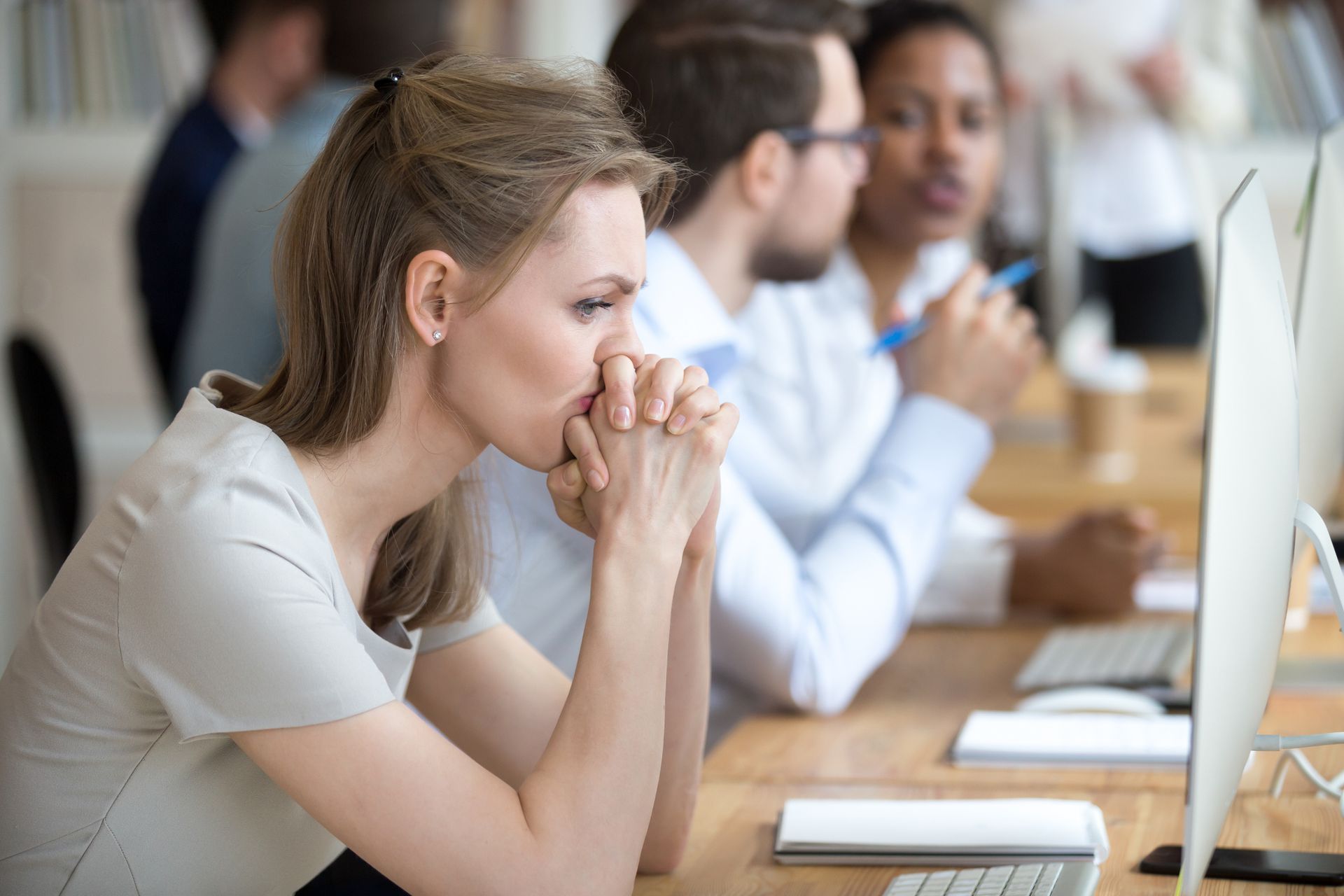 Woman with worried expression, hands clasped, looking at a computer screen in a busy office.