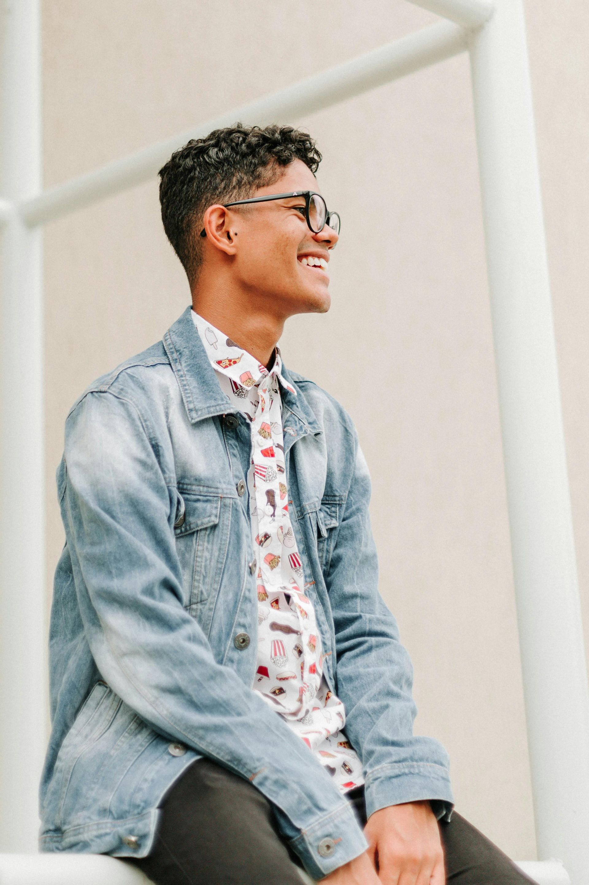 A young man wearing a denim jacket and glasses is sitting on a railing.