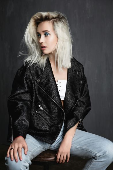 A woman is sitting on a stool wearing a black jacket and jeans.