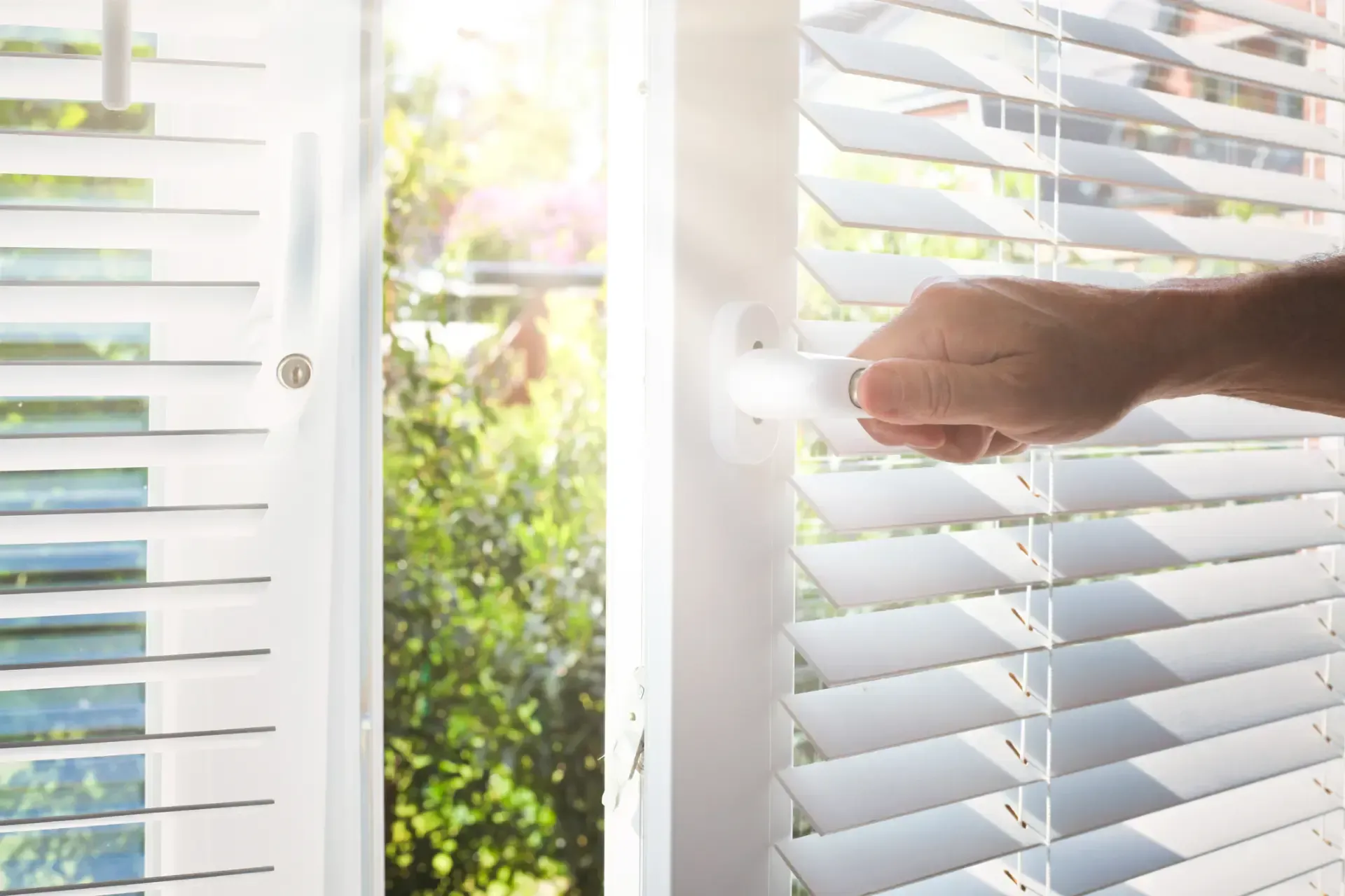 Finestra con persiane bianche che si aprono a mano, con luce solare e vegetazione sullo sfondo.