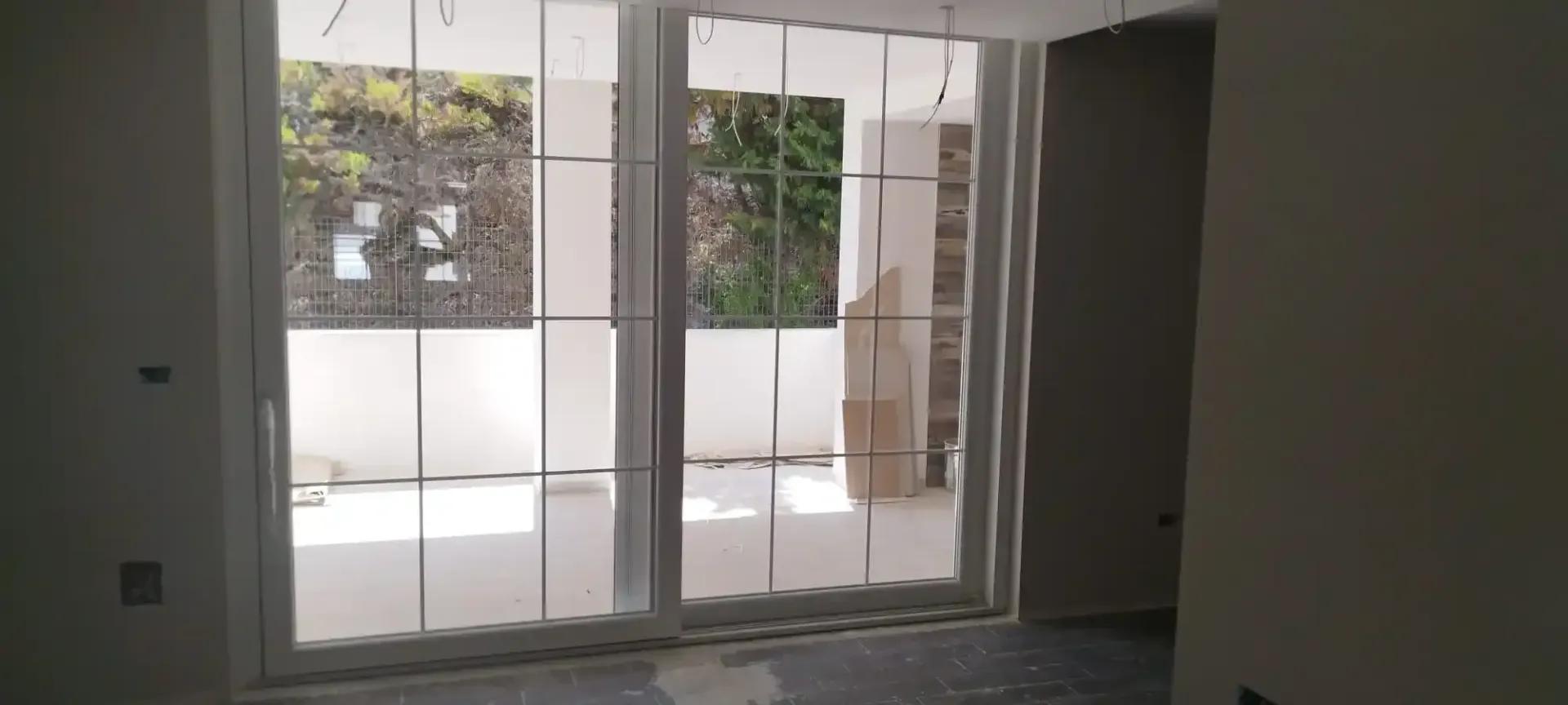 Stanza interna con grandi porte-finestre in vetro con cornice bianca che conducono a un balcone. All'esterno si vedono alberi e cielo.