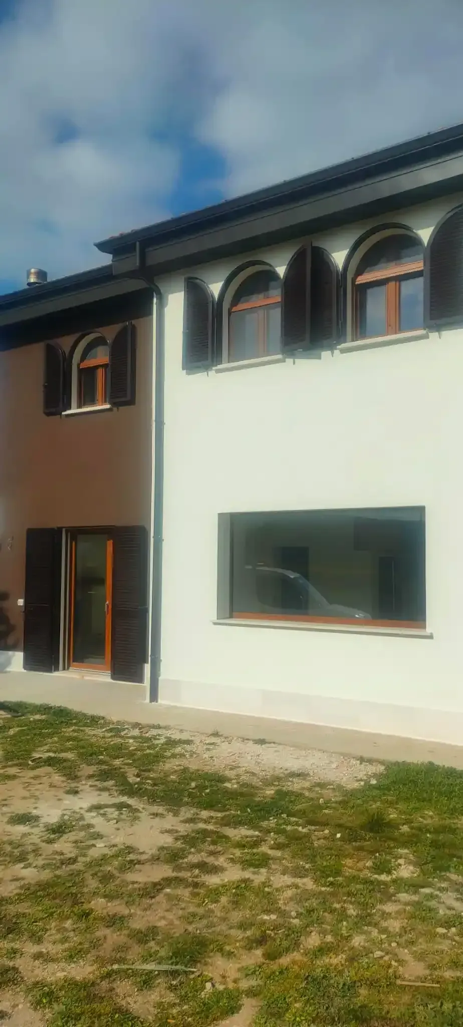 Esterno di un edificio a due piani con finestre ad arco, persiane scure e un prato verde sotto un cielo nuvoloso.