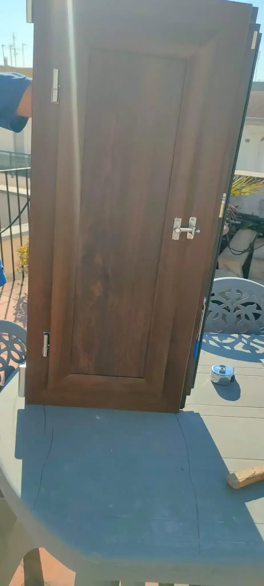 Una porta di legno invecchiato con ferramenta in metallo è appoggiata su un tavolo di plastica all'aperto. La luce del sole splende.