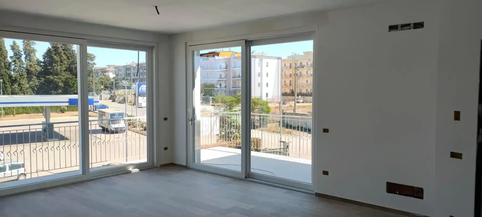 Interno di una stanza con ampie finestre che si affacciano su una strada cittadina. Le pareti sono bianche, il pavimento è di colore chiaro.