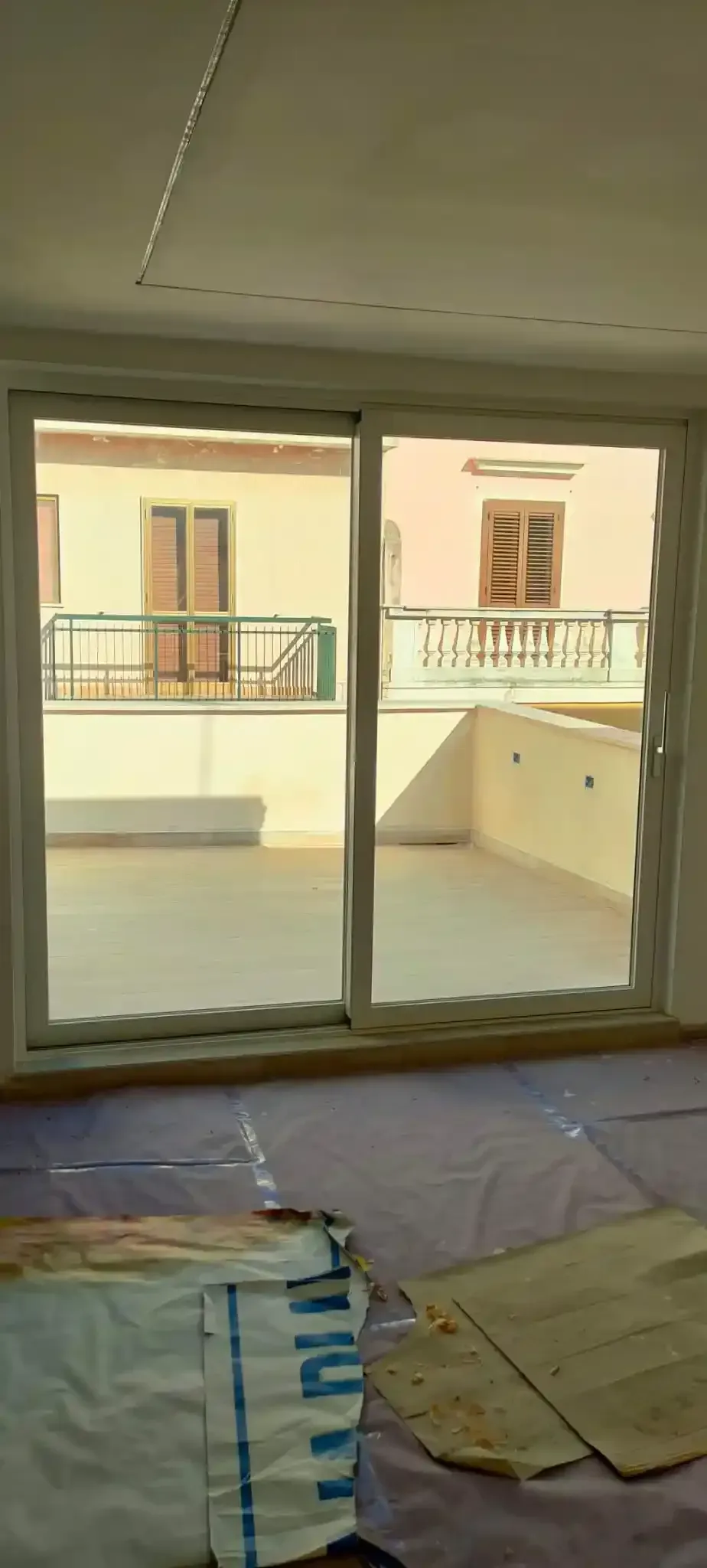 Vista interna: porte scorrevoli in vetro che si aprono su un balcone con altri edifici. Telo viola e detriti sul pavimento.