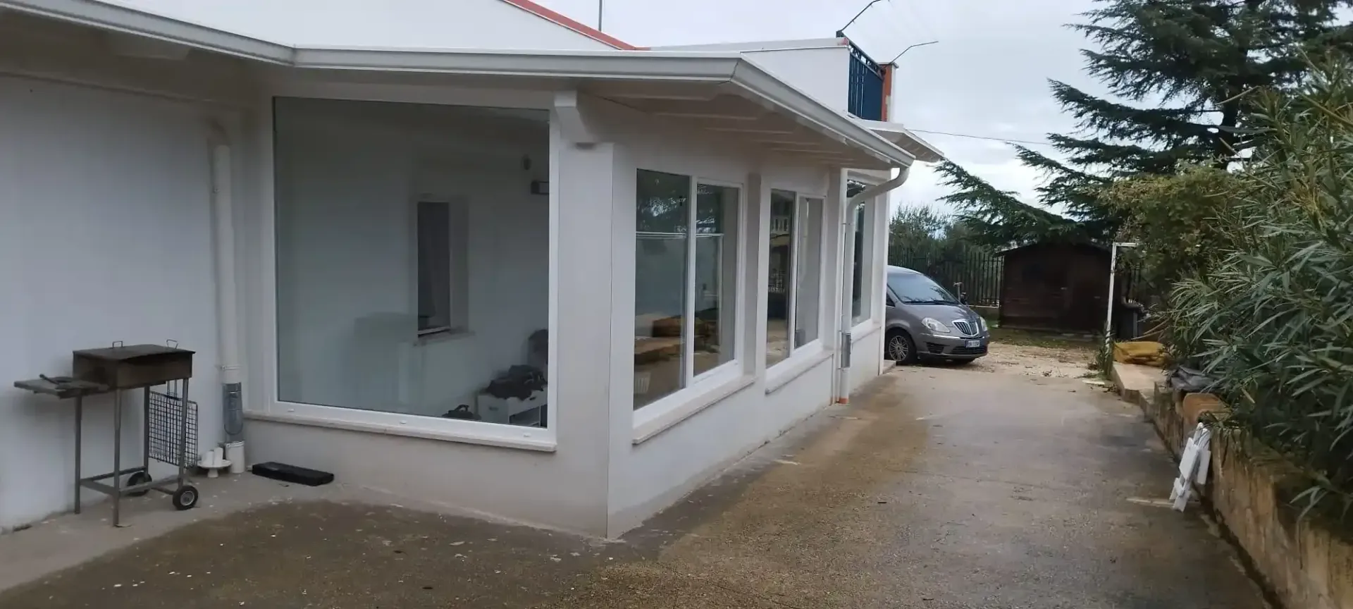 Vista esterna di un edificio bianco con ampie finestre. Un'auto è parcheggiata su un vialetto bagnato.