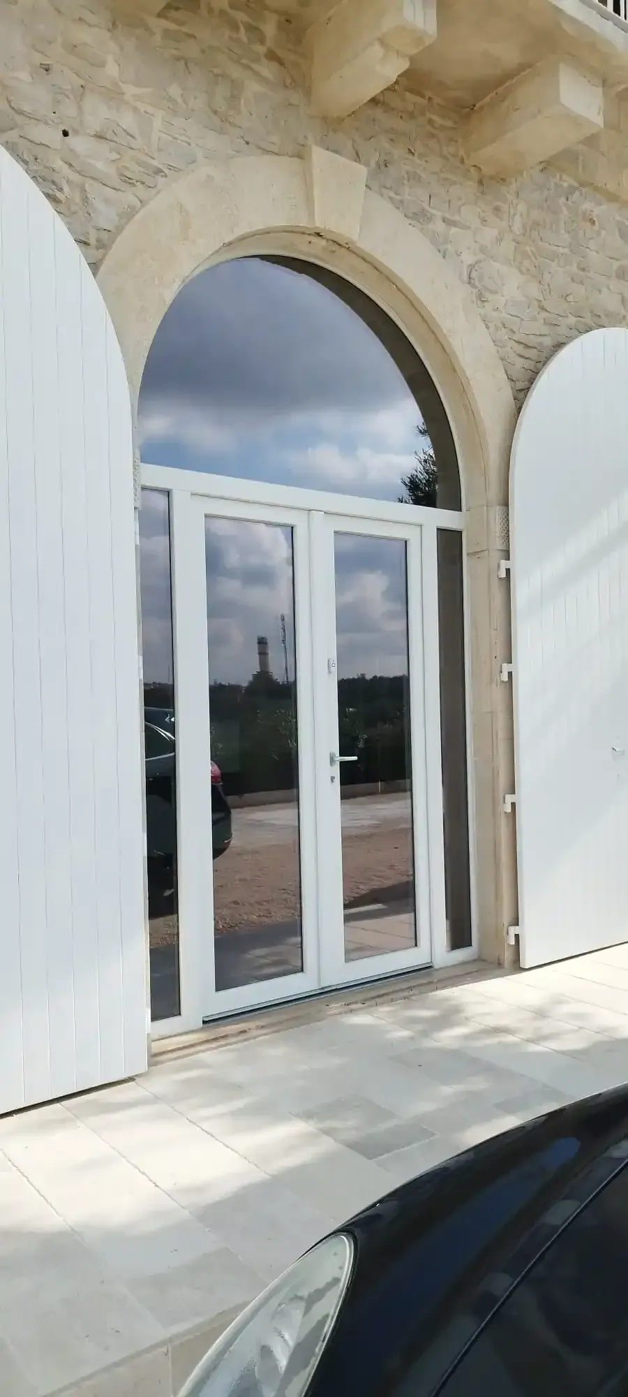 Porte in vetro con cornice bianca sotto un portale ad arco con persiane bianche e muro in pietra.