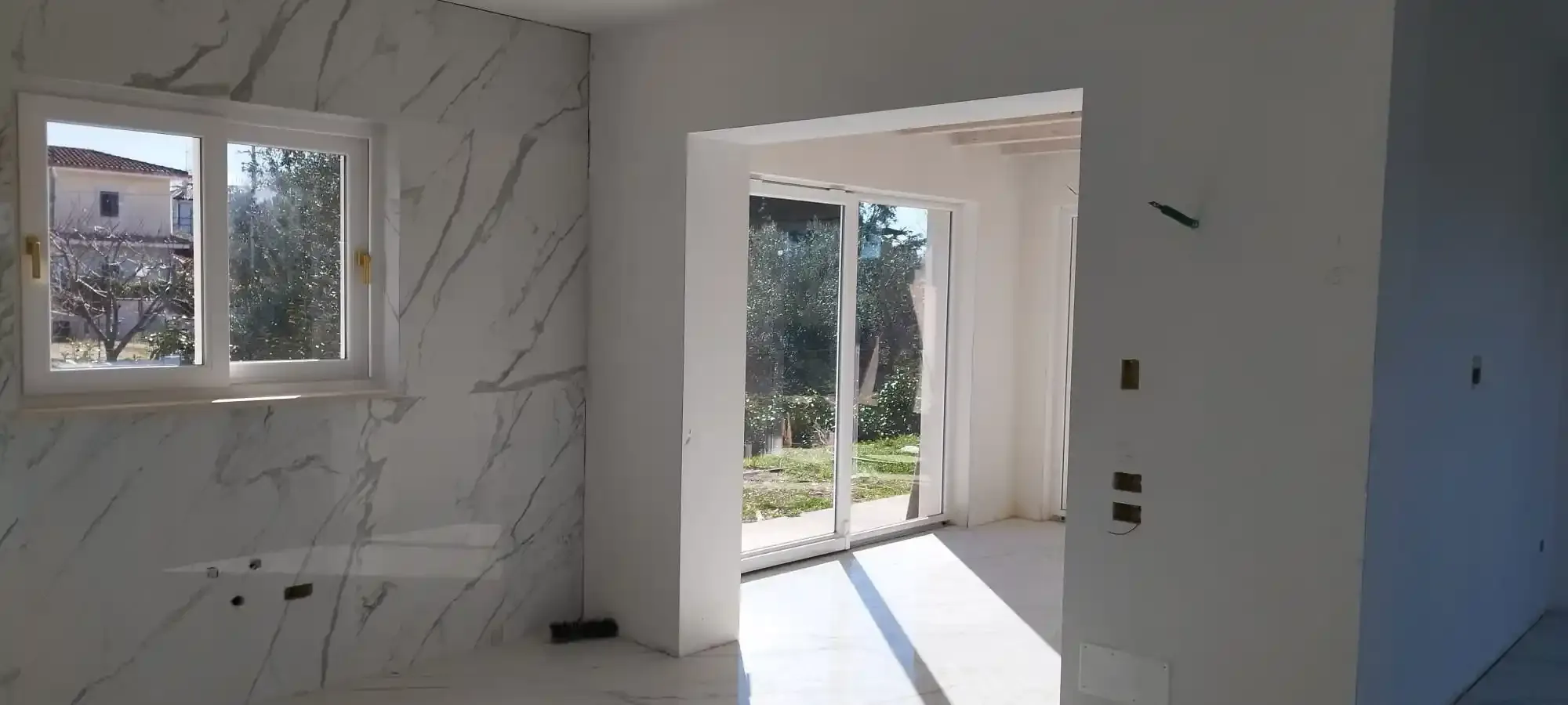 Vista interna di una stanza con parete in marmo, finestra e porta scorrevole in vetro. La luce del sole filtra attraverso le aperture.