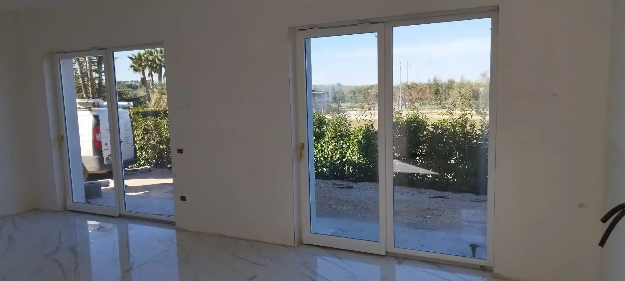 Vista interna di una stanza con due grandi porte a vetri, pareti bianche e vista sul cortile.
