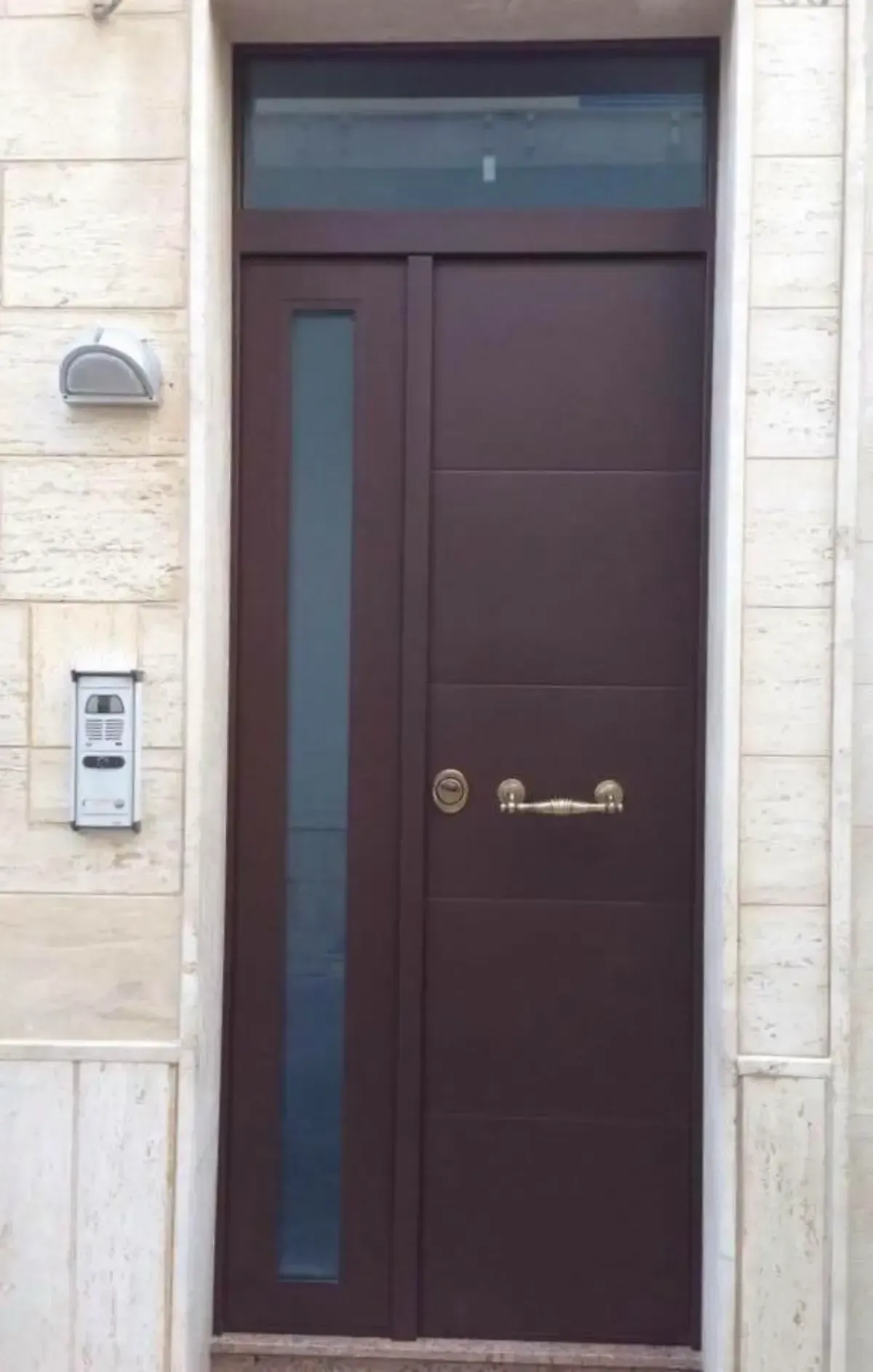 Porta d'ingresso marrone con finestra laterale e sopraluce, maniglia in ottone, incastonata in pietra color crema.