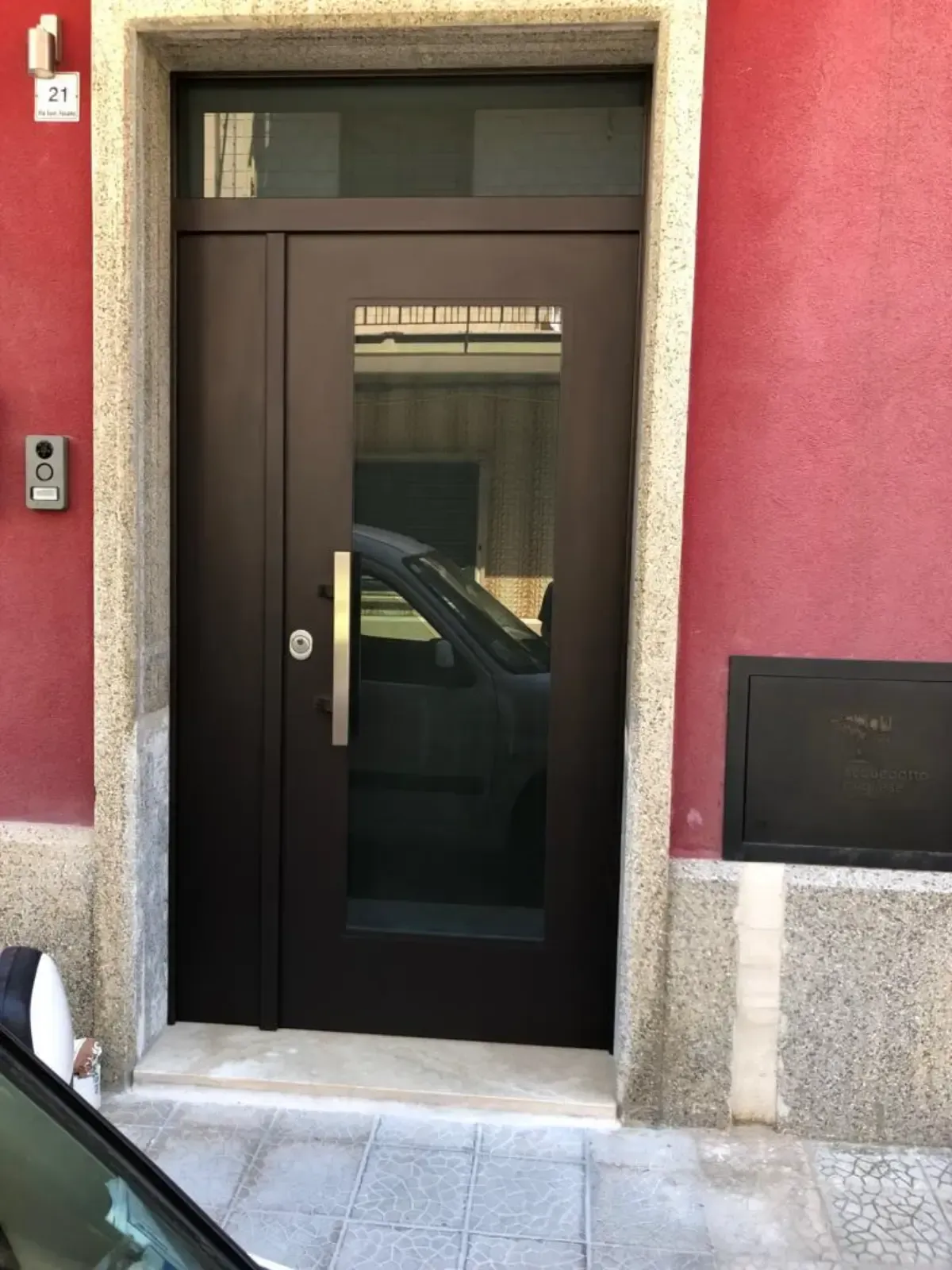 Porta marrone con finestra, maniglia argentata, inserita in una parete rossa con finiture in cemento.