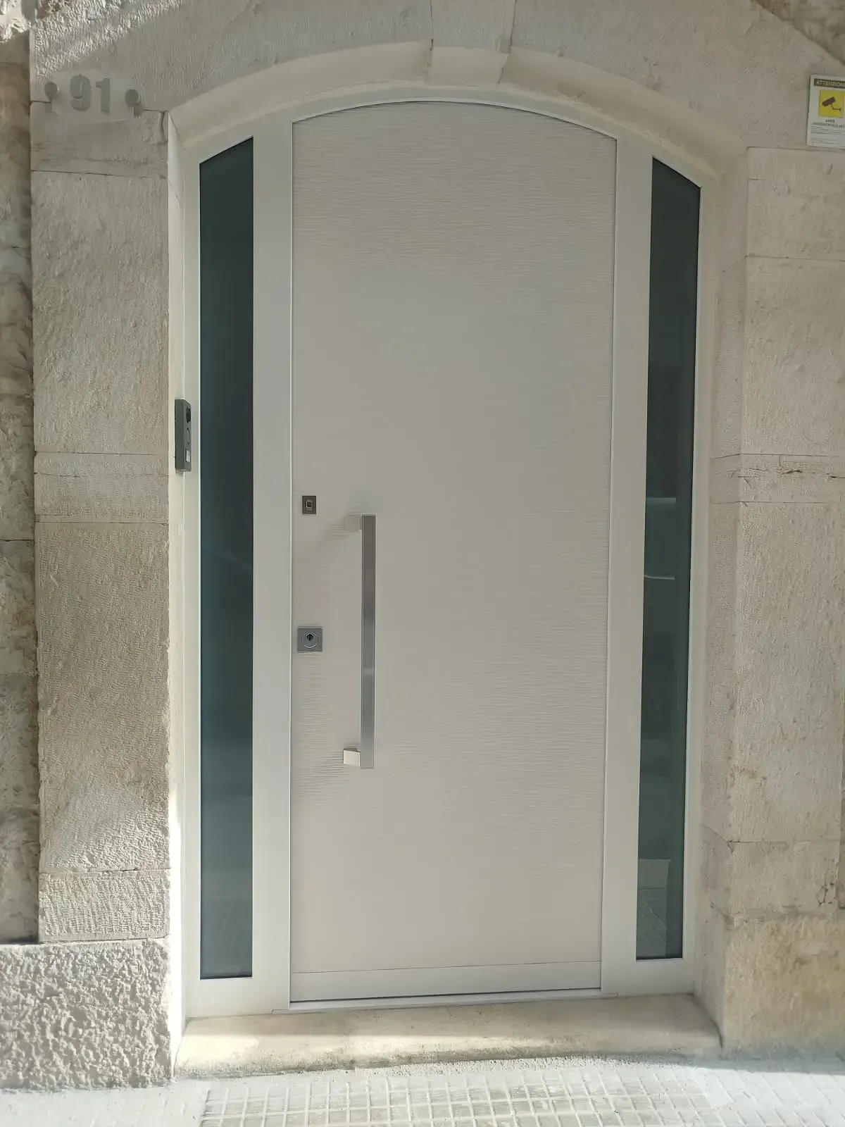Porta d'ingresso bianca con lunga maniglia argentata e pannelli laterali in vetro, incastonata in un edificio in pietra.
