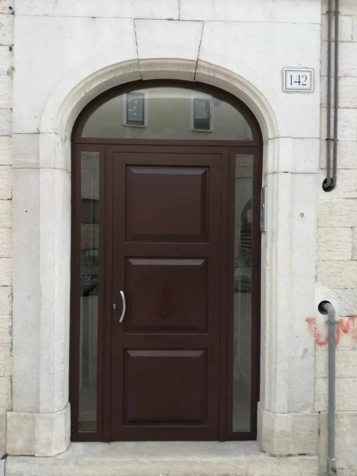 Porta ad arco marrone con finestre laterali, inserita in un portale in pietra bianca, numero 142 sopra.