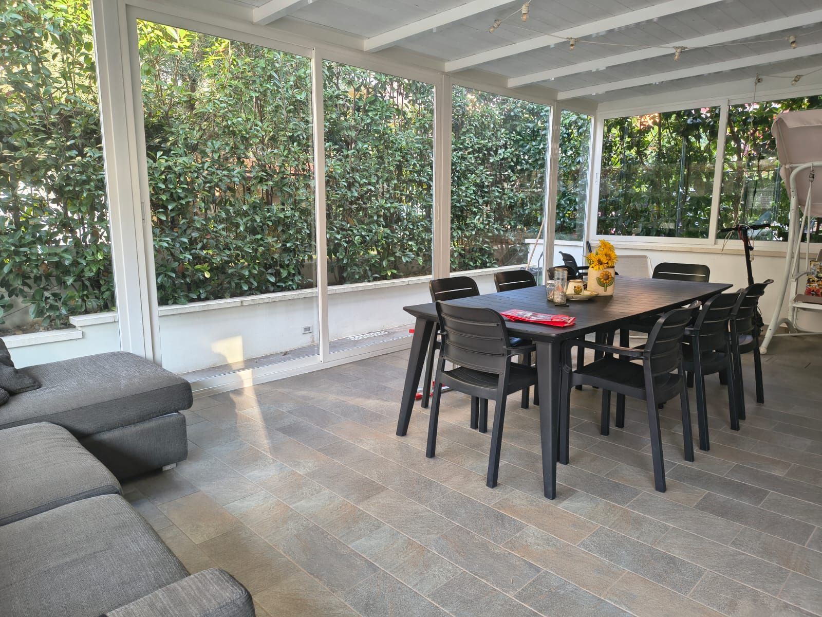 Zona pranzo esterna con vetrate, tavolo apparecchiato per sei persone e vista su un prato verde e alberi.