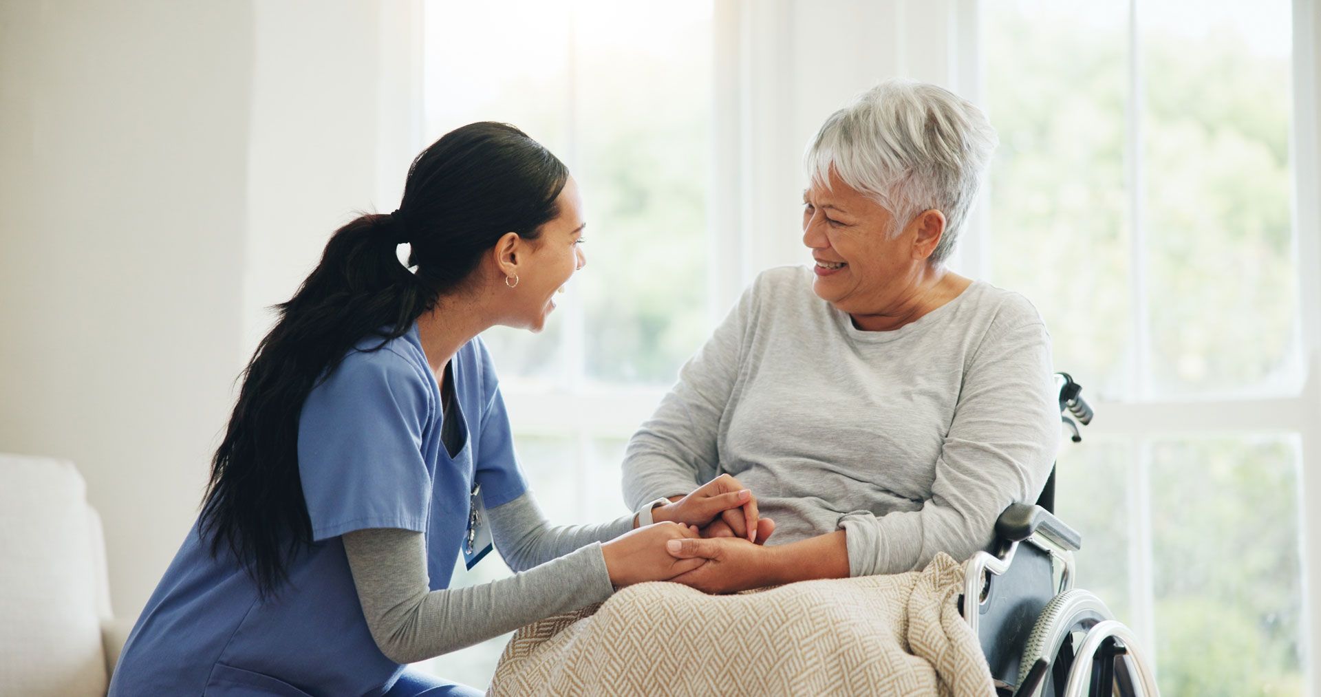 A smiling caregiver in scrubs kneels to hold the hands of a person sitting in a wheelchair, fostering a warm connection.