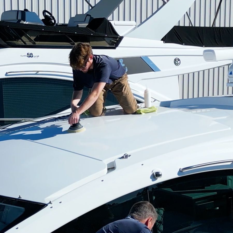 Man kneeling on a white boat deck, polishing it with a machine. Another man works below.