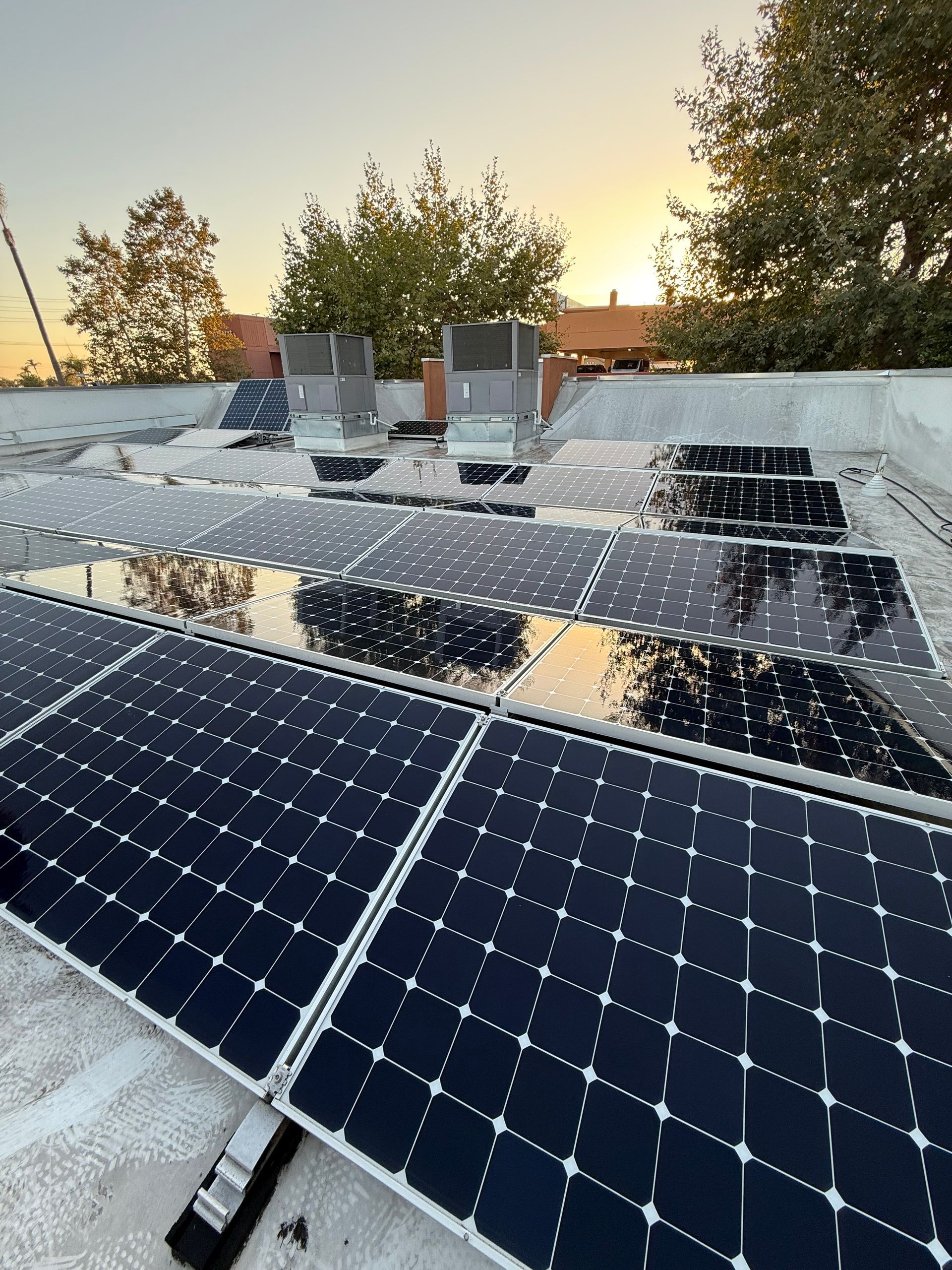 Solar panels installed on a flat roof reflect a colorful sunset. Air conditioning units are also present.