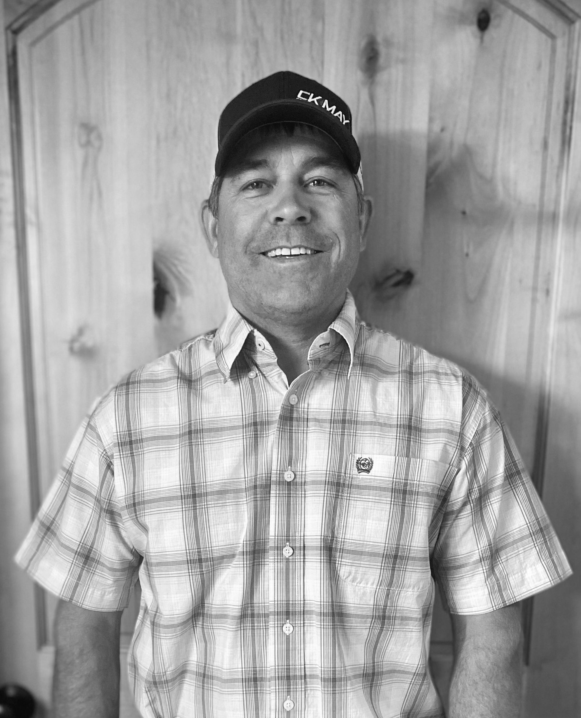 A smiling person wearing a baseball cap and a short-sleeved plaid button-down shirt, standing in front of a wood door.