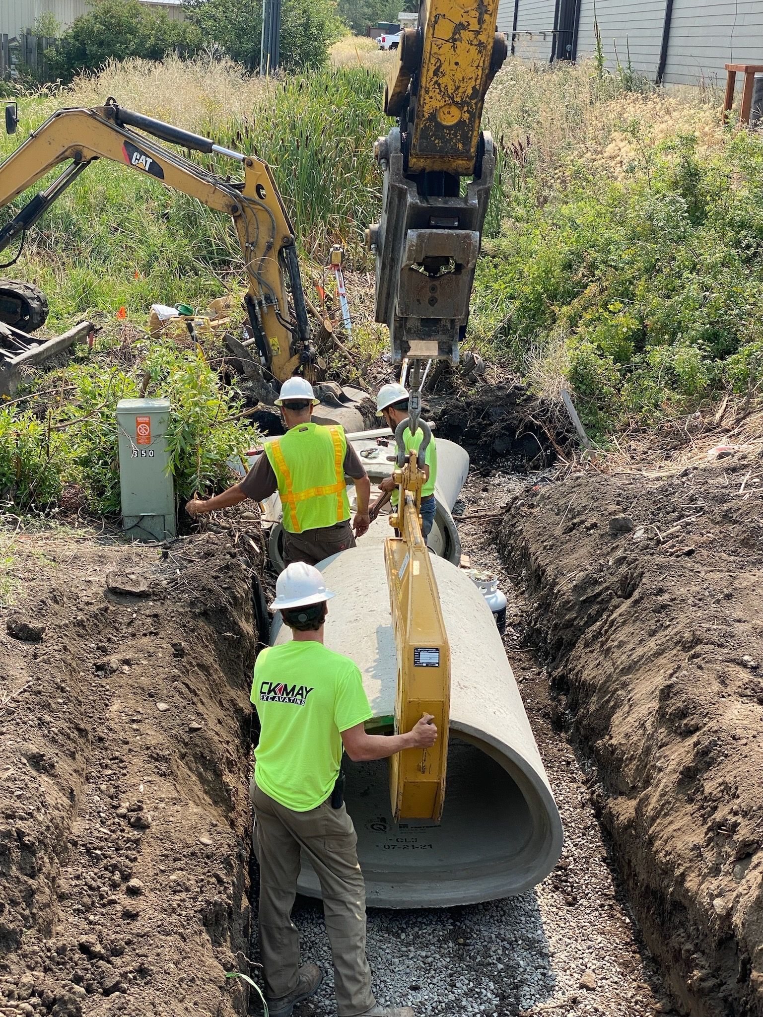 Two construction workers in high-visibility gear guide a concrete pipe segment being lowered into a trench by an excavator.