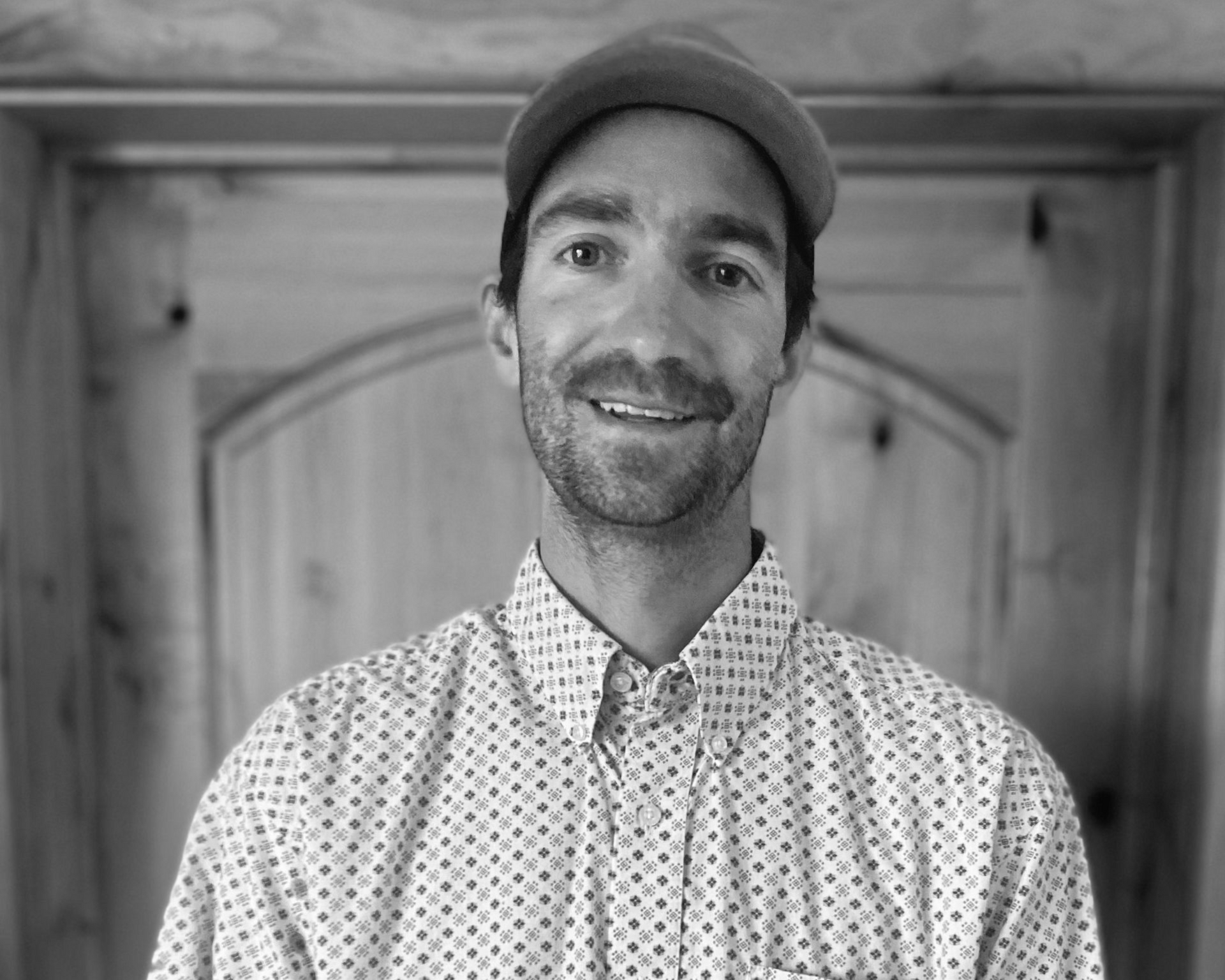 A black-and-white portrait of a smiling person wearing a baseball cap and a patterned button-down shirt against a door.