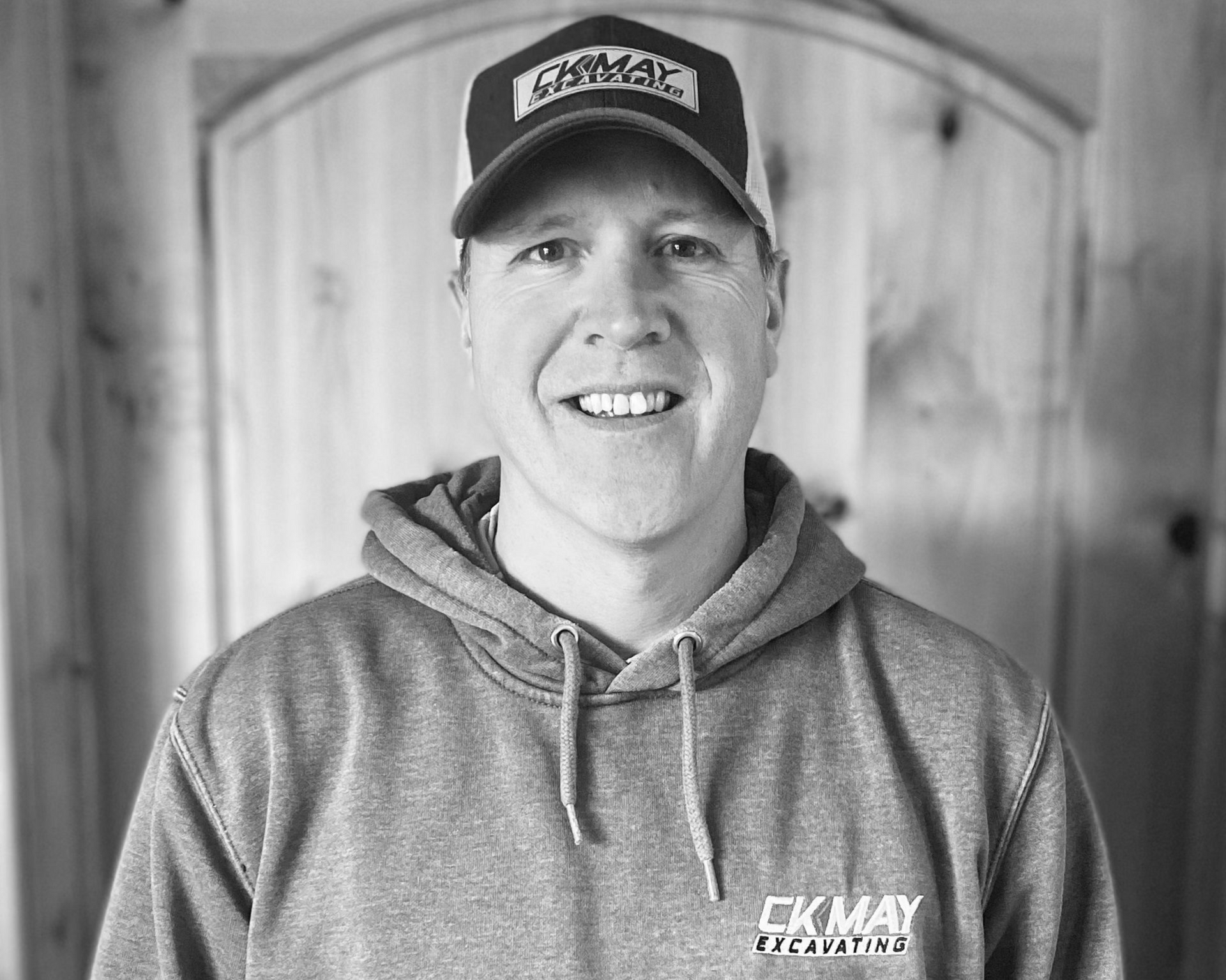 A person wearing a hooded sweatshirt and cap with logos, smiling in front of a wood-paneled door in black and white.
