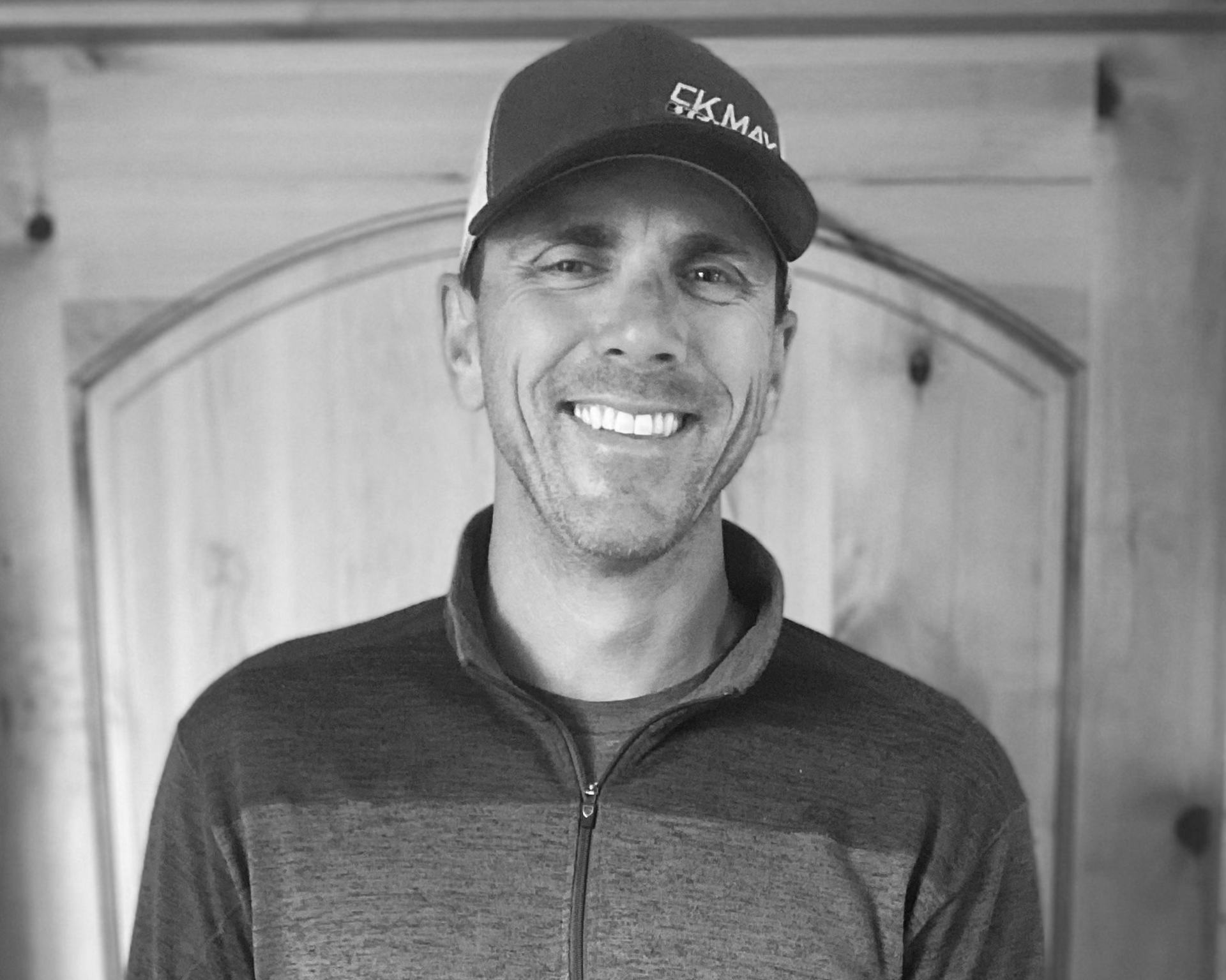 A smiling person wears a baseball cap and a zip-up jacket against a light wood-paneled background.