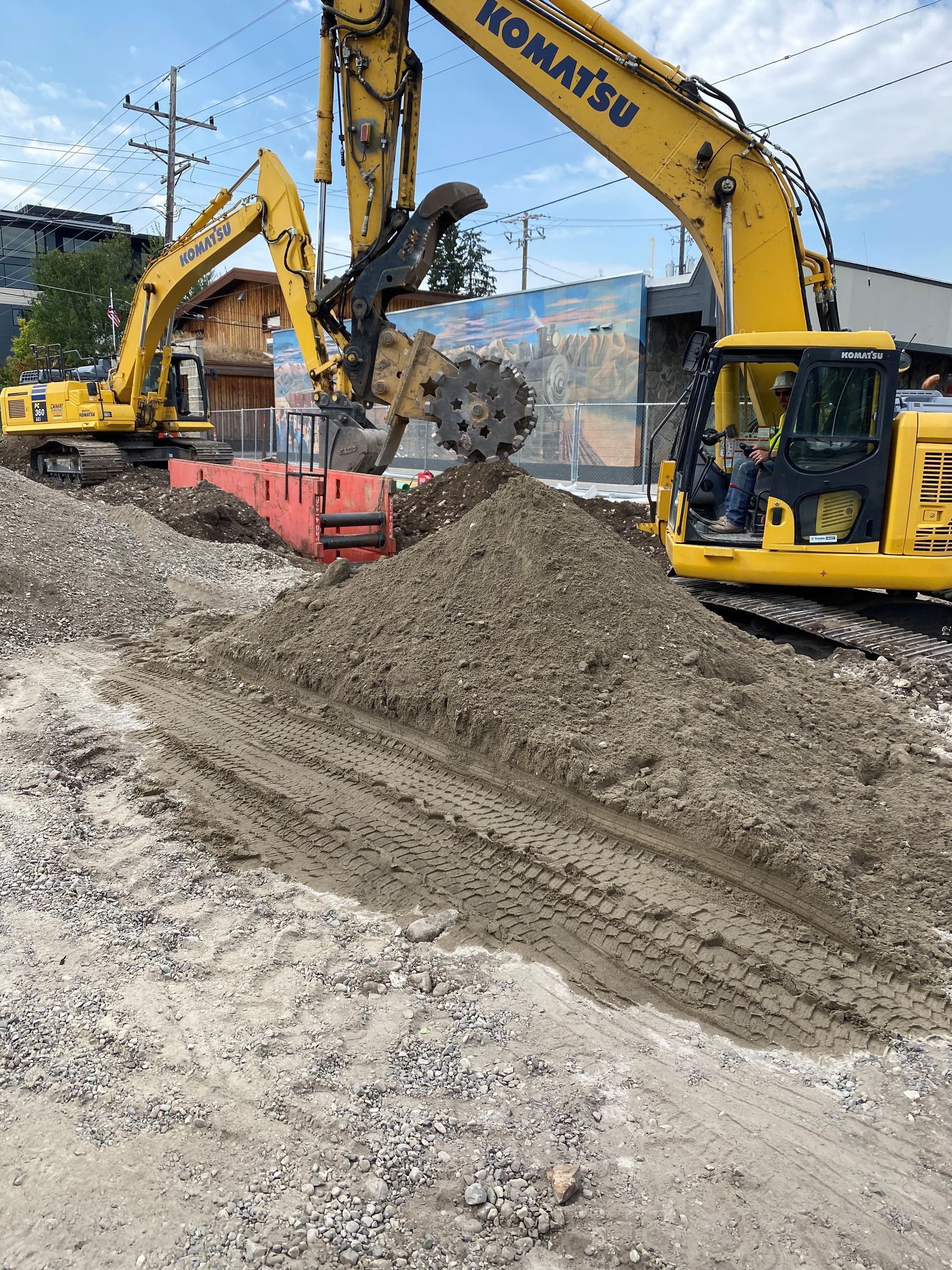 Two yellow Komatsu excavators operate at a construction site with piles of dirt, gravel, and a red concrete barrier.