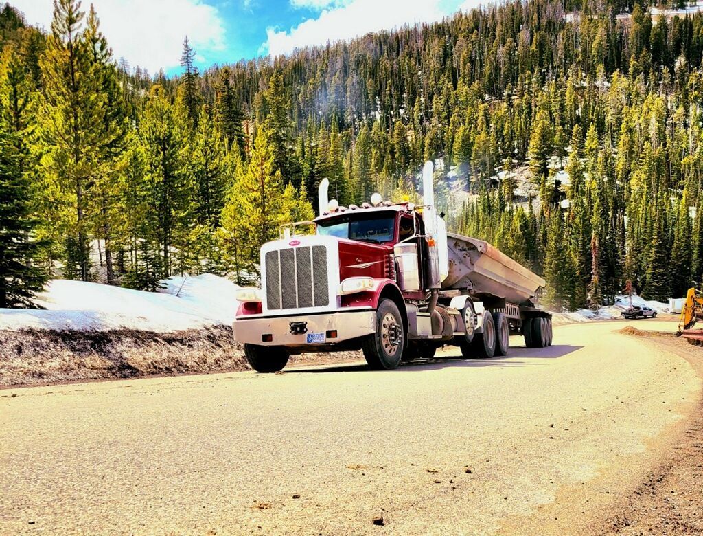 A maroon semi-truck pulls a flatbed trailer down a dirt mountain road lined with pine trees and patches of snow.
