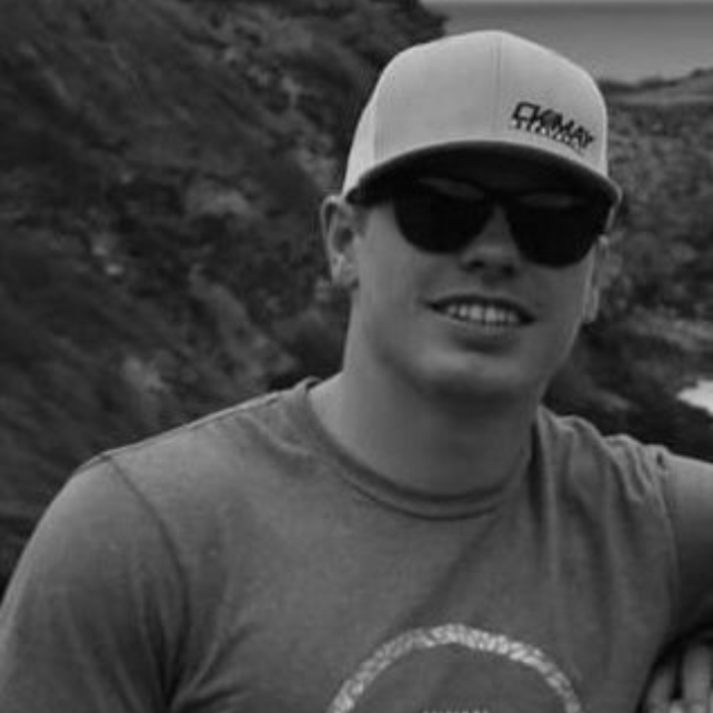 A person wearing a baseball cap and dark sunglasses smiling, positioned outdoors against a rocky landscape.