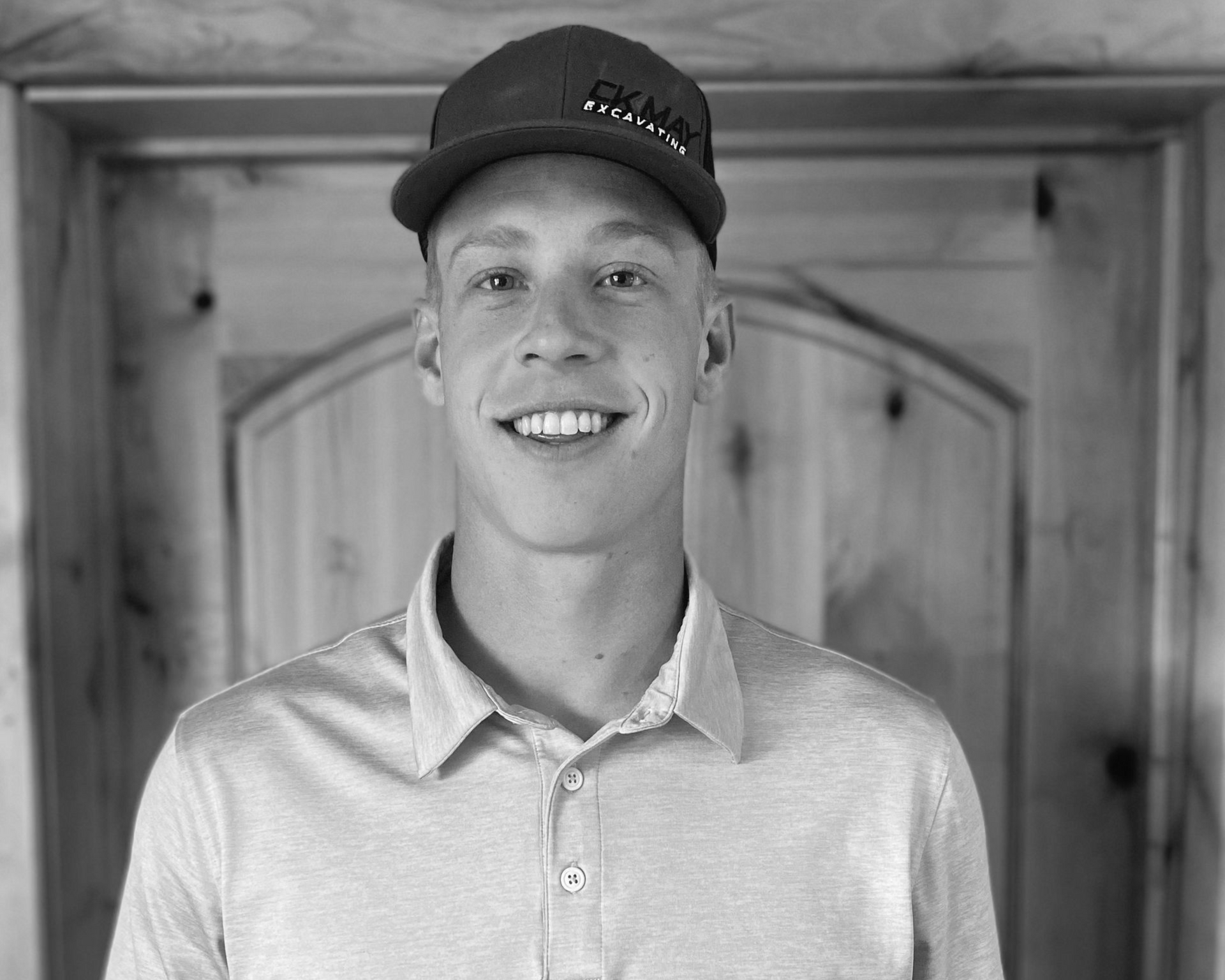 A smiling individual wearing a baseball cap and a polo shirt, centered in front of a wooden door.
