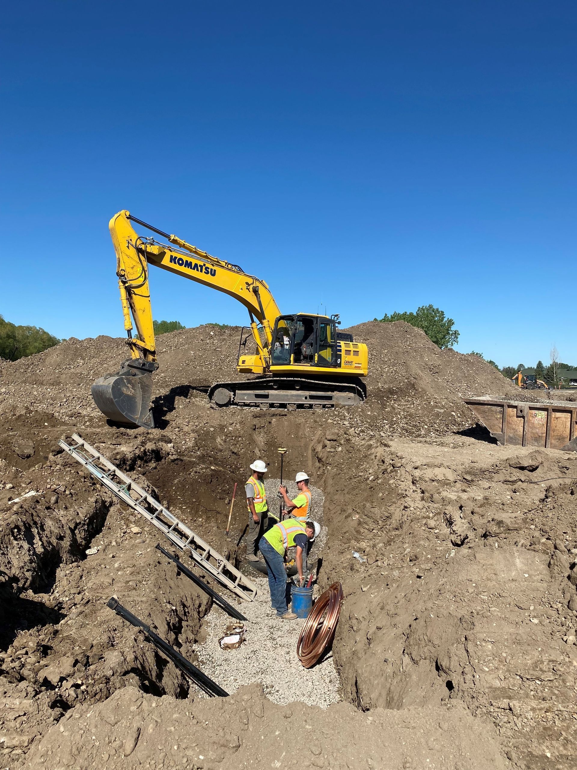 Construction workers in high-visibility vests work in a dirt trench near a yellow Komatsu excavator under a blue sky.