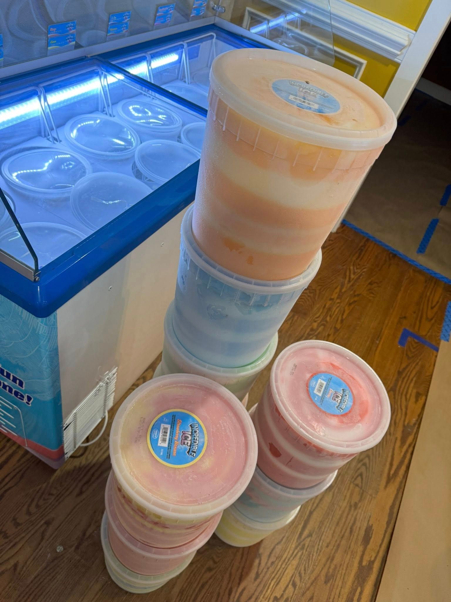 A stack of tall orange and blue cotton candy tubs next to smaller pink and white tubs near a refrigerated display case.