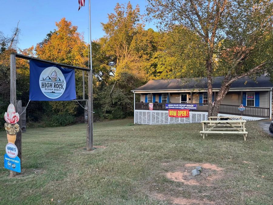 A grassy yard with an ice cream cone sign, a blue flag, and a single-story building with a porch and picnic table.