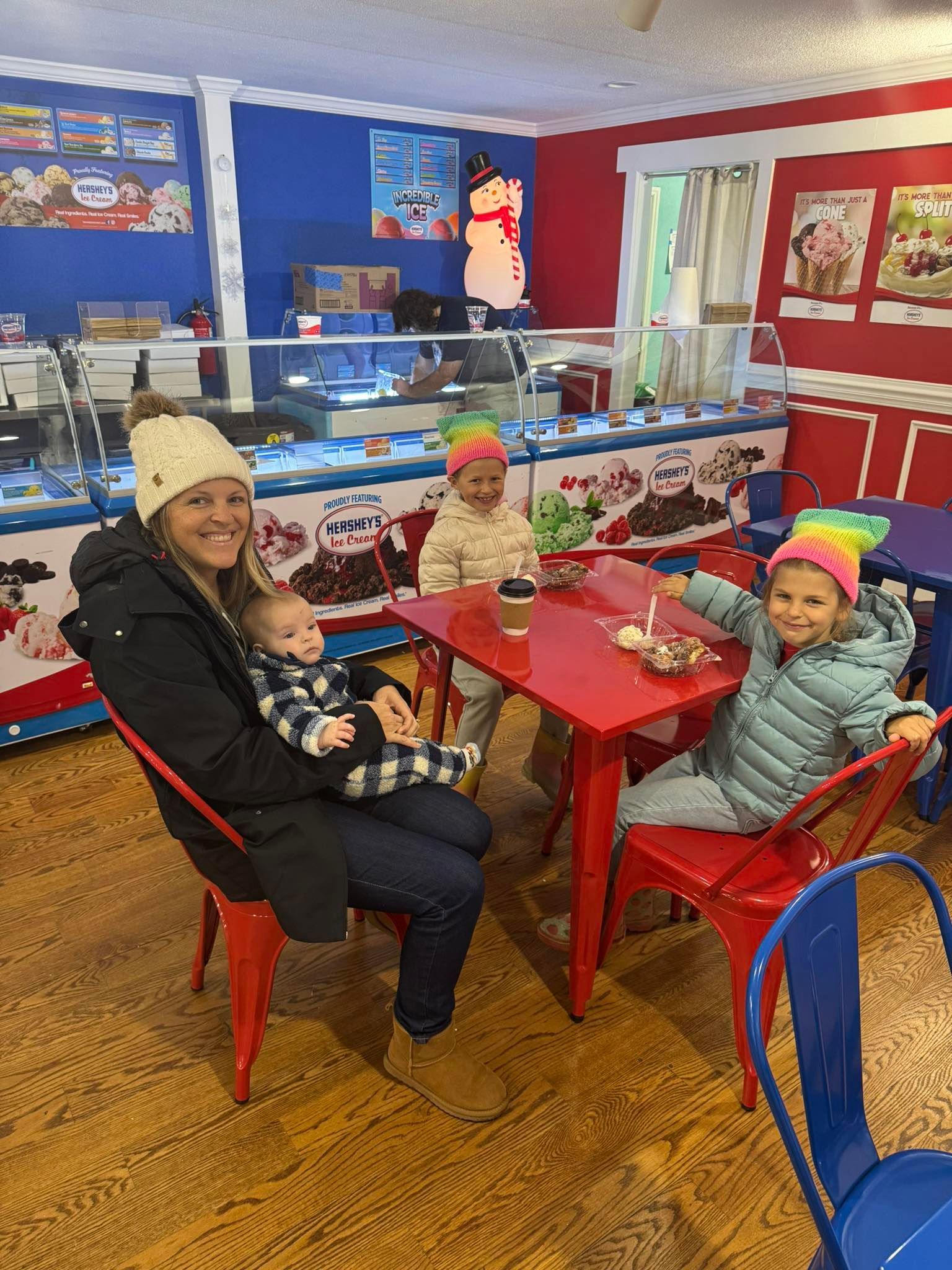 A smiling person holding a baby, sitting at a red table with two other children in an ice cream shop with blue walls.