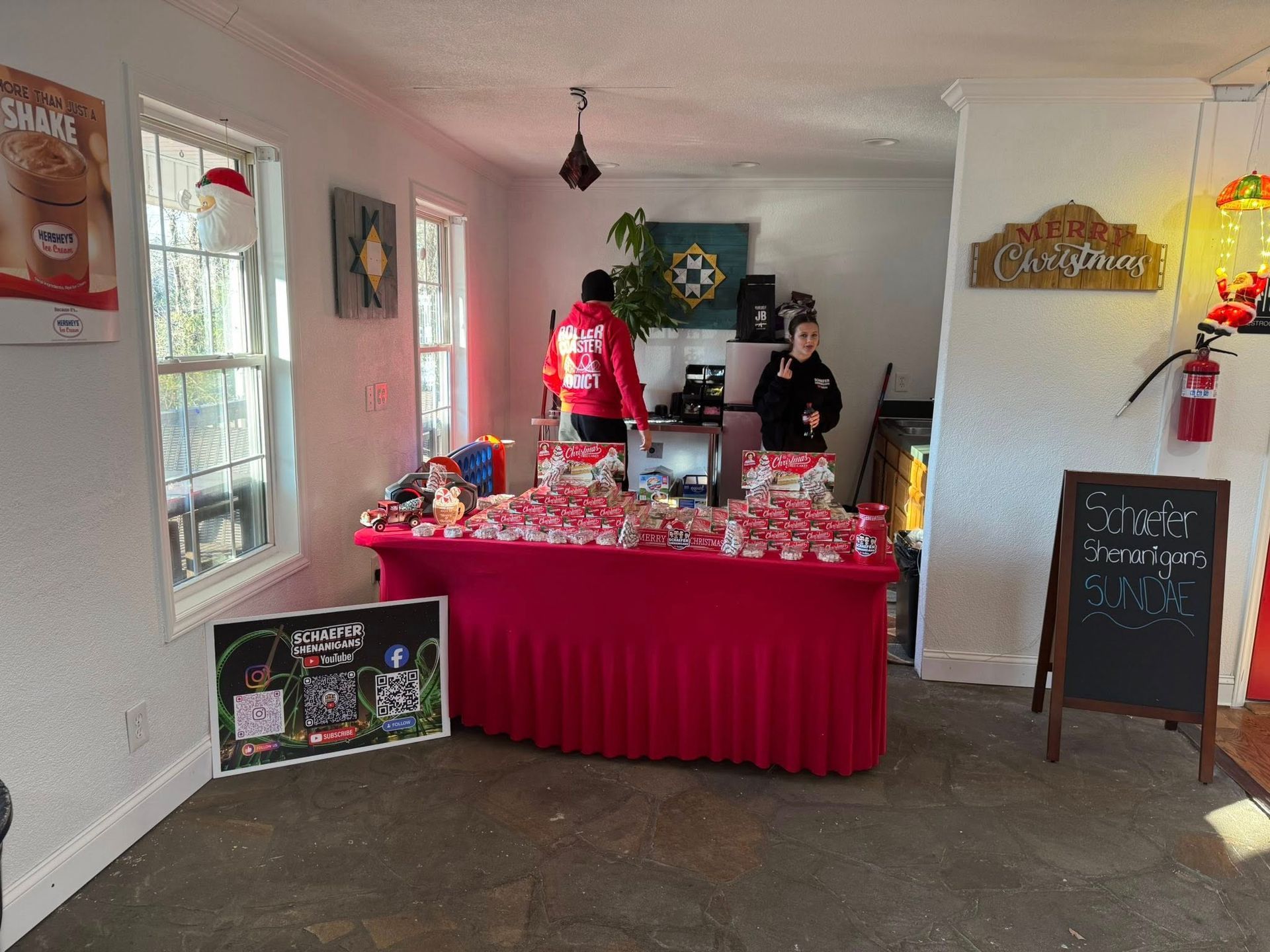 Two people stand behind a red-clothed table covered in small red items in a shop, with signage on the wall and floor.