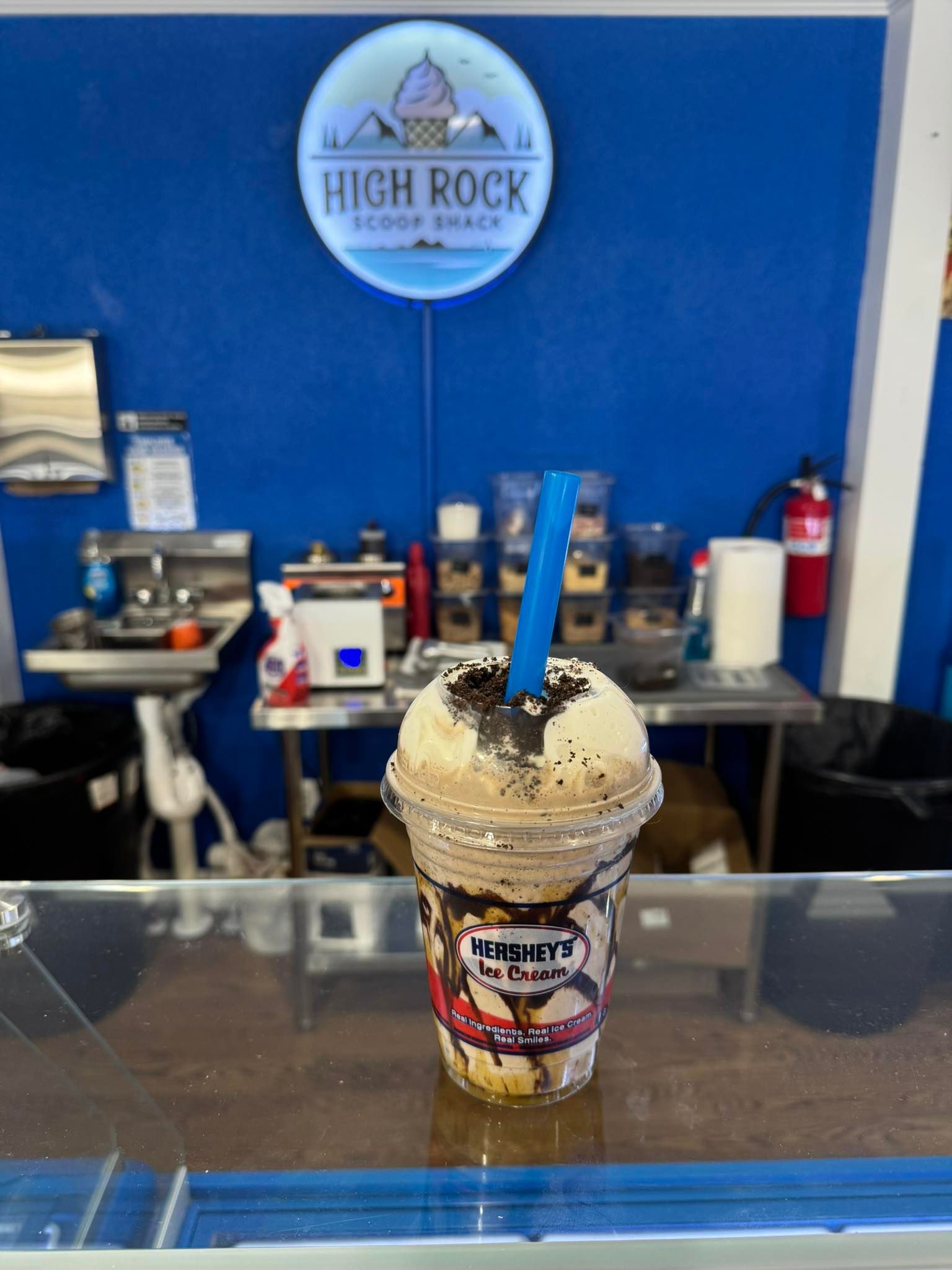 A chocolate milkshake with a blue straw sits on a counter in front of the High Rock Scoop Shack sign and prep area.