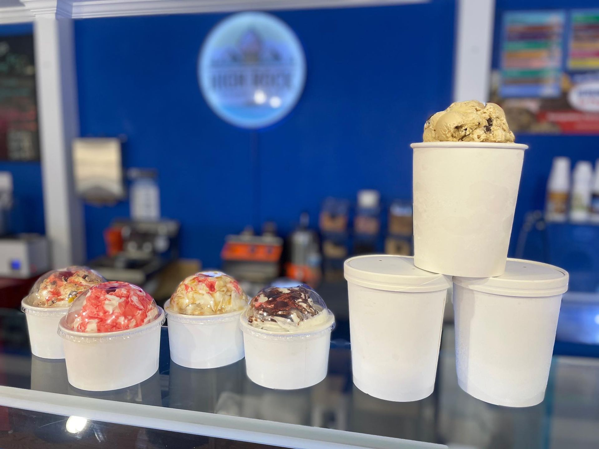 Several ice cream cups with various flavors lined up on a counter in front of a blue wall in an ice cream shop.