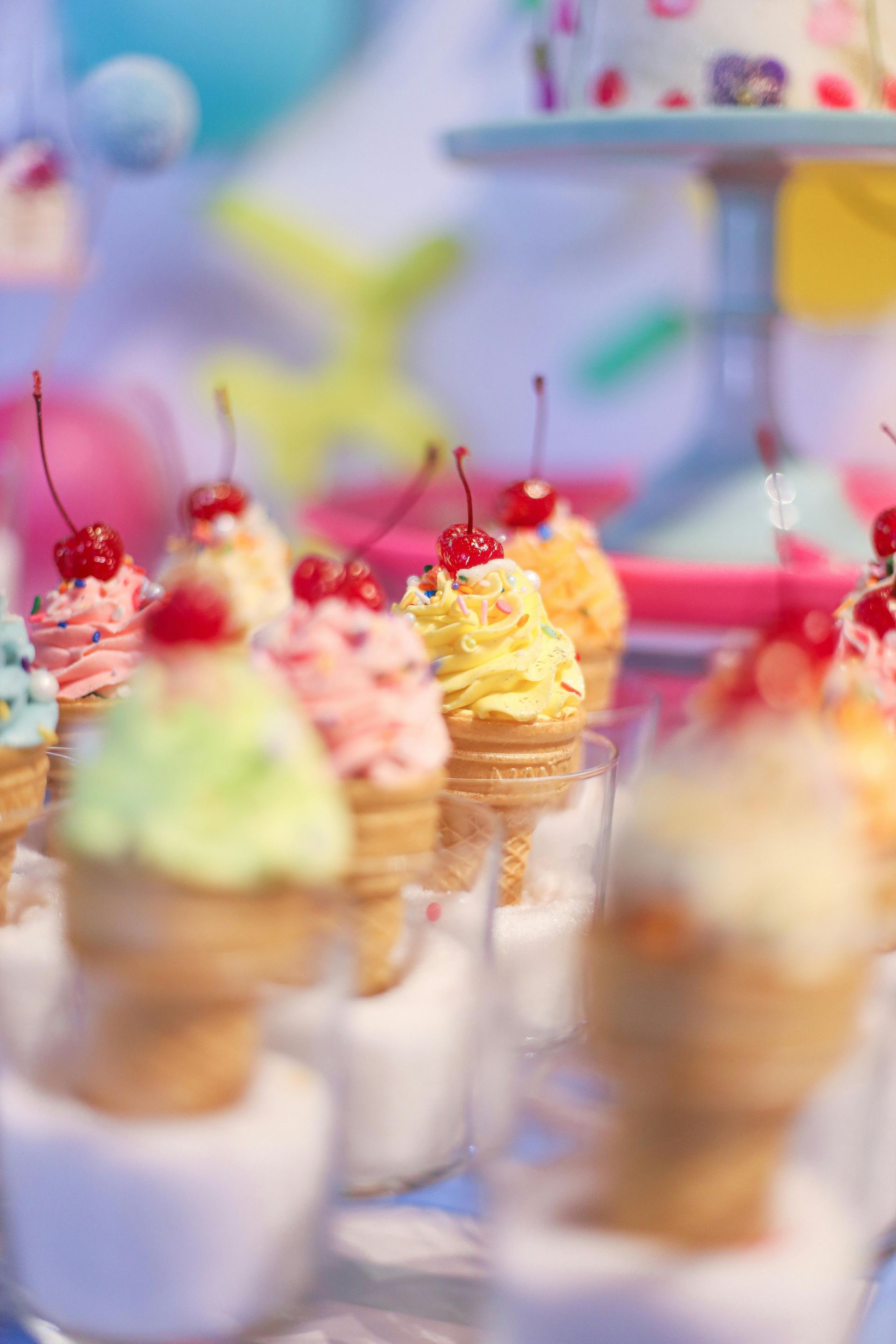 A collection of mini ice cream cones topped with sprinkles and cherries, standing in clear cups at a party.