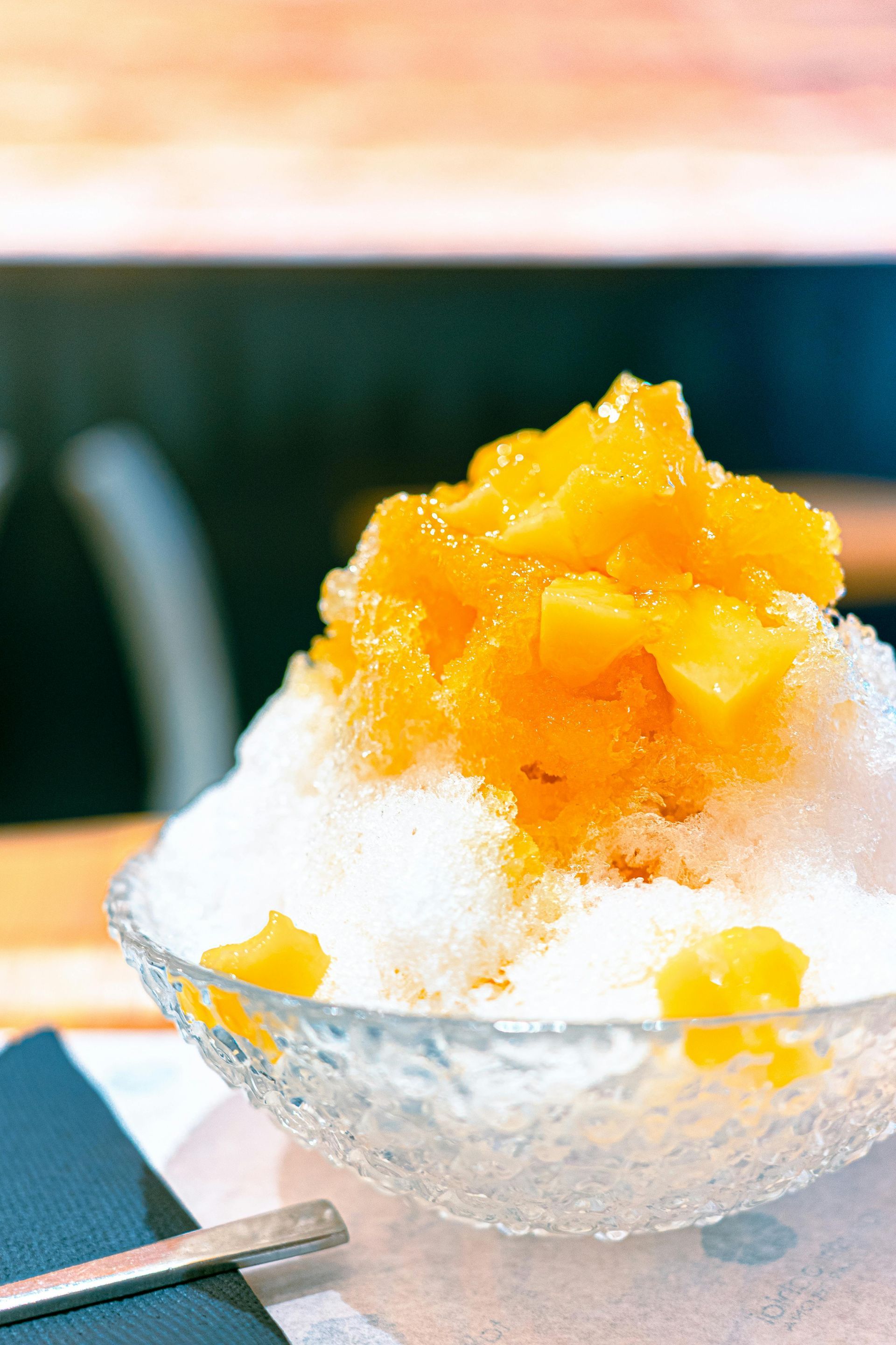 A crystal bowl filled with fluffy shaved ice topped with bright orange diced mango chunks on a table.