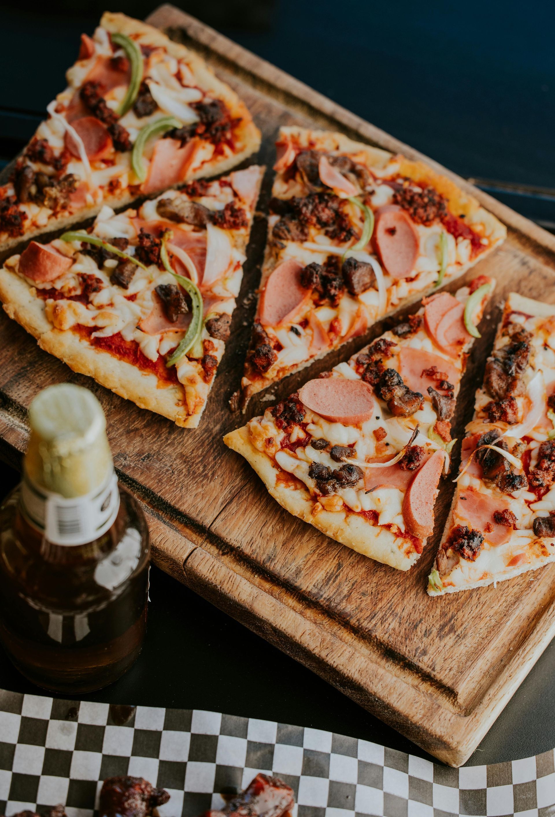 Four pizza slices with sausage and peppers on a wooden serving board next to a bottle of beer.