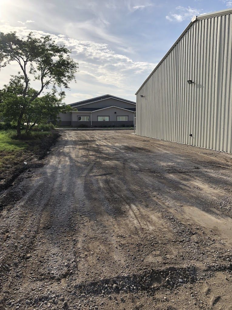 A dirt road leading to a building with a warehouse in the background.